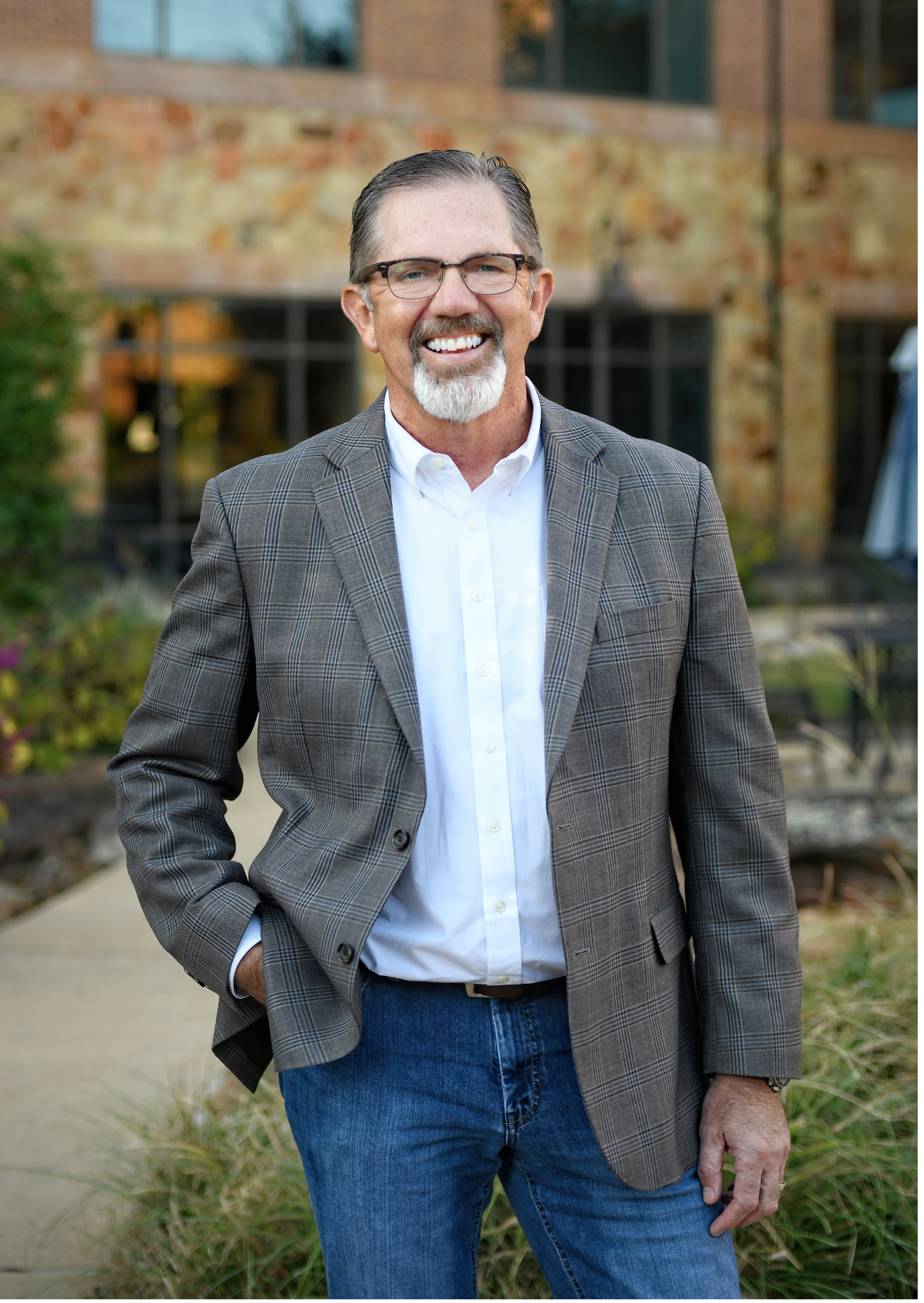 A middle-aged man with gray hair, beard, and glasses, dressed in a white shirt and plaid blazer, standing outdoors in front of a modern brick building with windows and greenery.