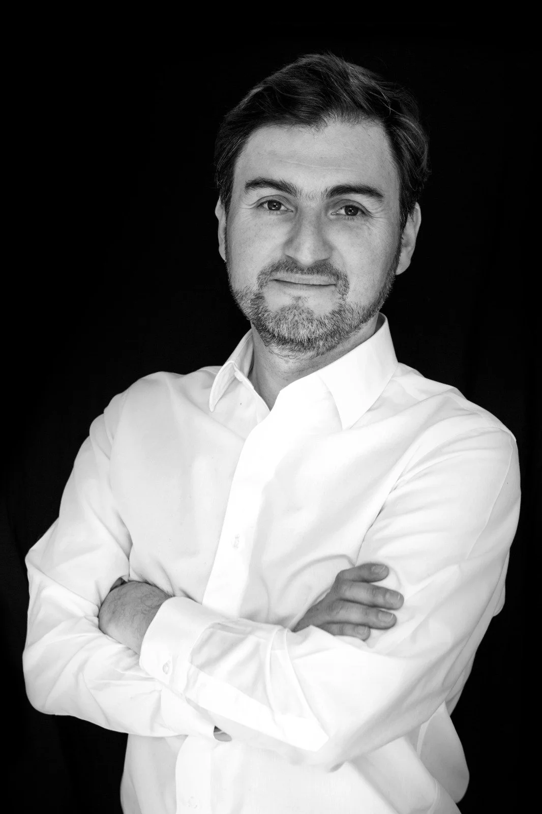 Retrato en blanco y negro de un hombre con barba y cabello oscuro, vistiendo camisa blanca, con brazos cruzados y sonriendo ligeramente, fondo negro.