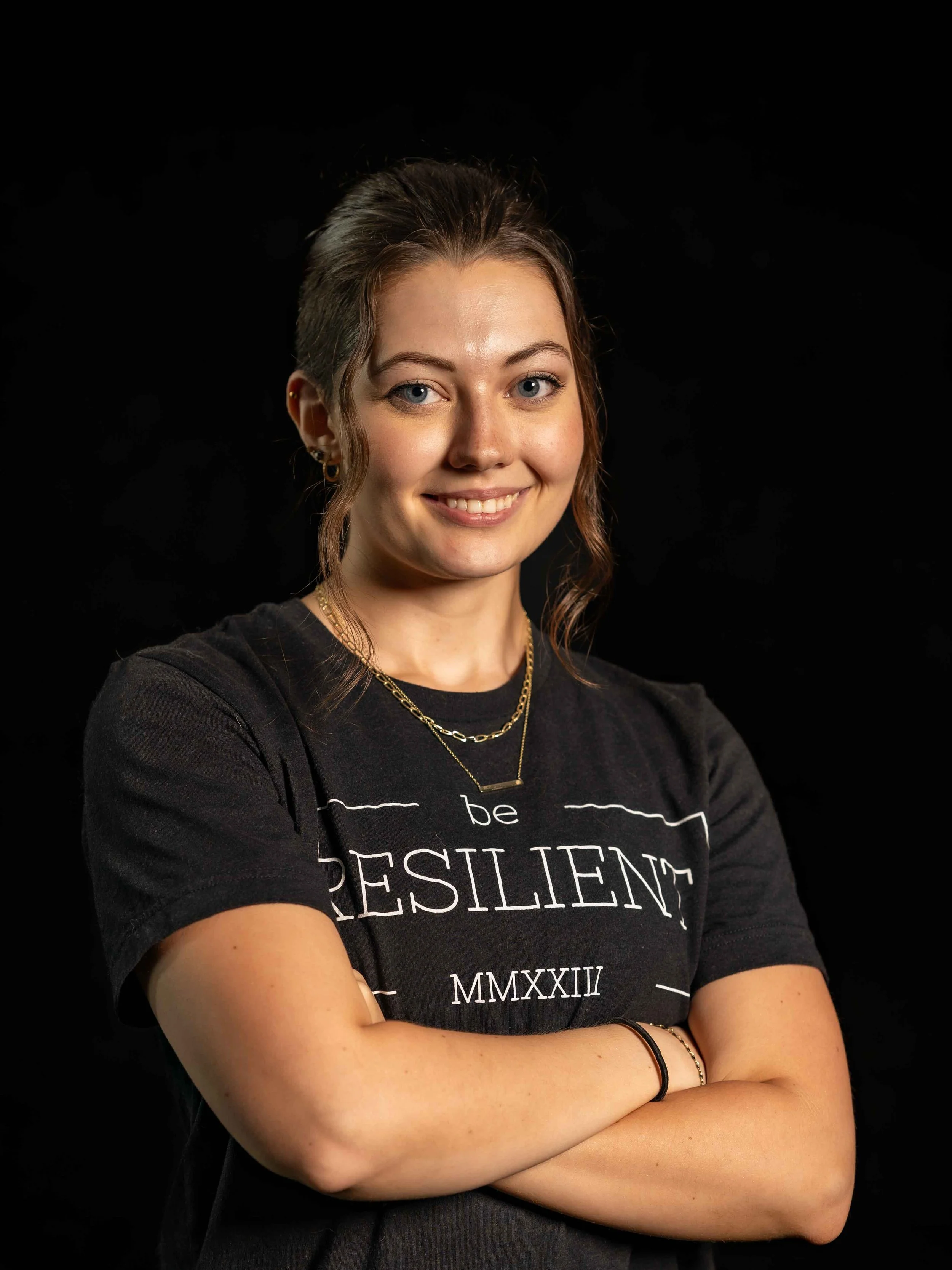 Portrait of a young woman with light skin, blue eyes, and brown hair, smiling with arms crossed, wearing a black T-shirt with white text and jewelry, against a dark background.