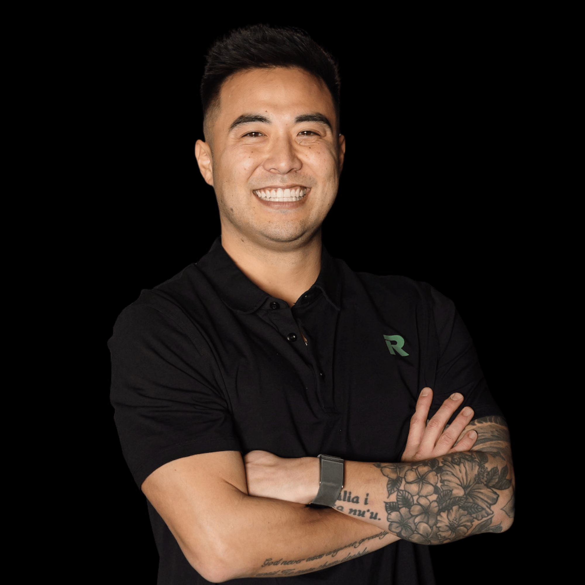 Smiling man with crossed arms, wearing a black polo shirt, tattoos on left arm, and a fitness tracker on right wrist, against a black background.