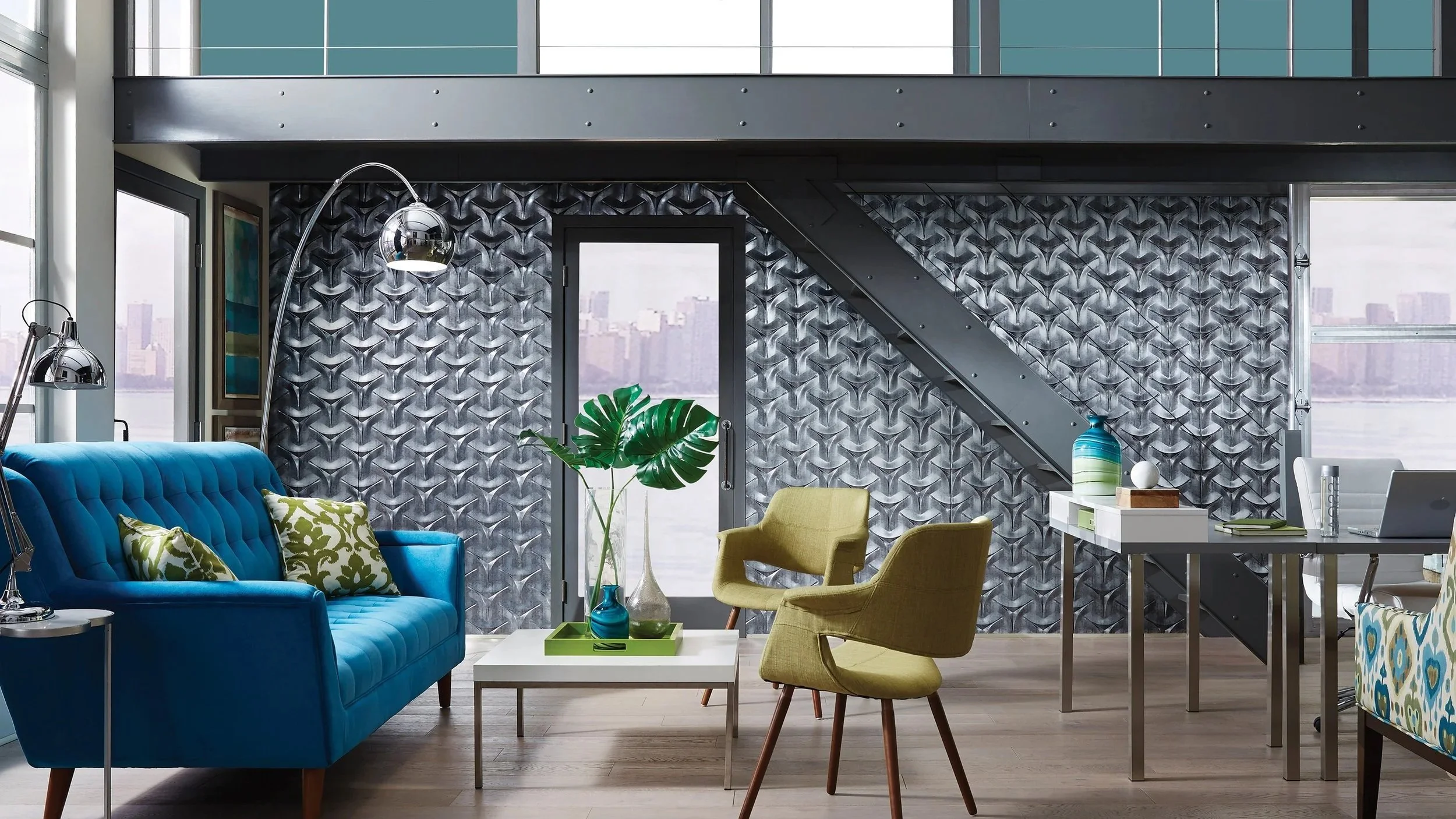 Modern living room with a blue velvet sofa, green armchair, and yellow armchair. Decorative pillows on the sofa, large windows with city view, patterned black and silver accent wall, greenery in vases, and a white desk with decorative objects.
