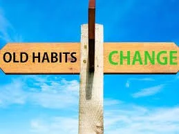 A wooden signpost with two arrows, one saying 'OLD HABITS' pointing left, and the other saying 'CHANGE' pointing right, against a bright blue sky with clouds.