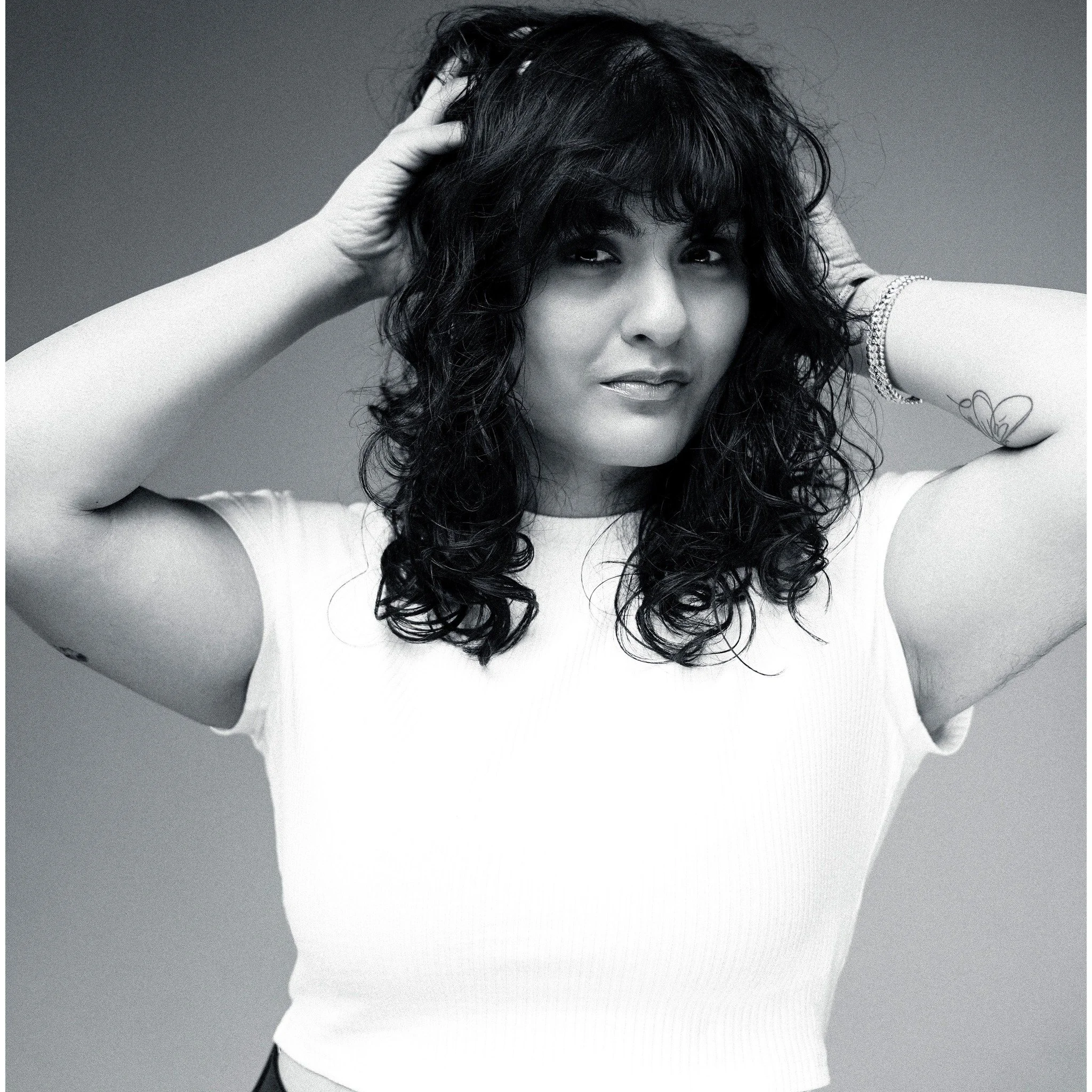 A woman with curly dark hair, wearing a white top and a bracelet, is looking at the camera while touching her hair with both hands. The image is in black and white.