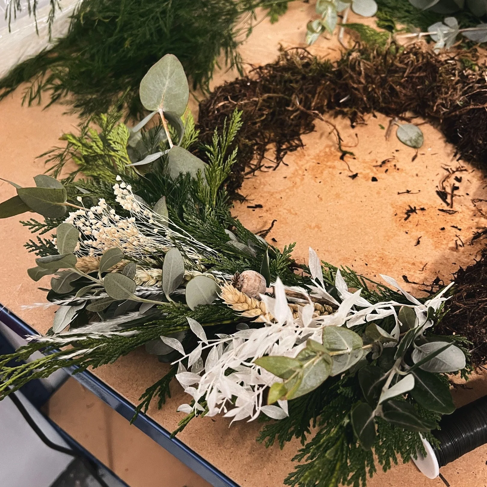 A wreath of green and white foliage with eucalyptus leaves, pine, and dried flowers on a wooden table.