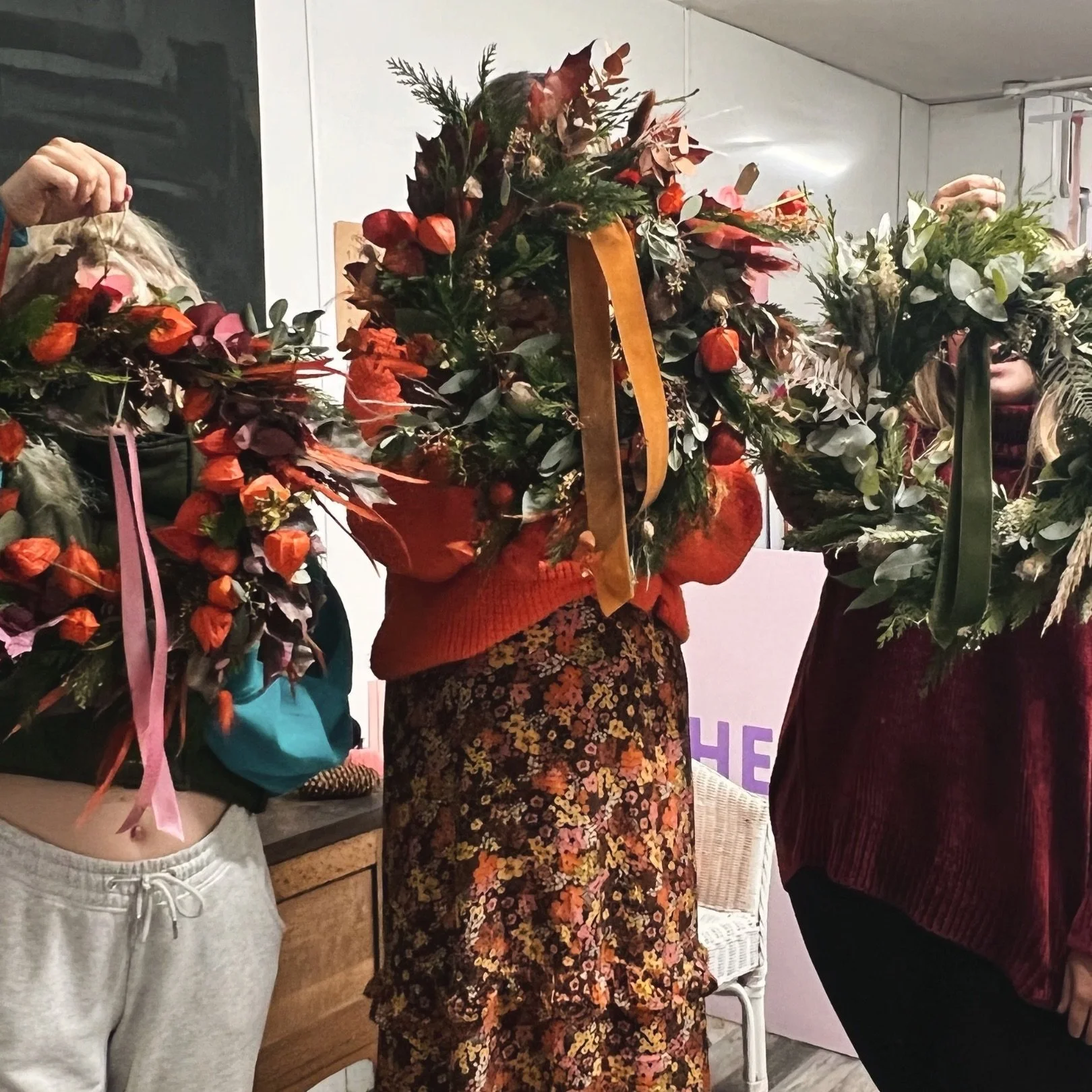 Three women holding large floral wreaths, partially obscuring their faces, in an indoor setting.