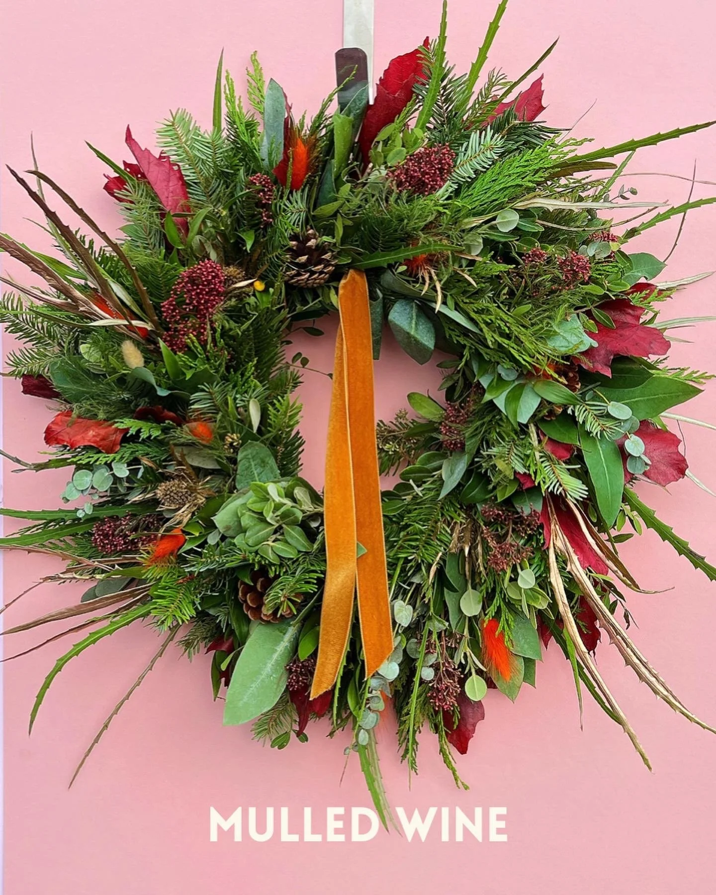 A festive holiday wreath with greenery, pinecones, red and green leaves, and orange accents hanging on a pink background, with the words 'MULLED WINE' at the bottom.