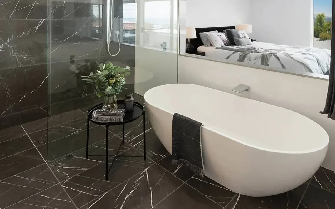 Modern bathroom with a black marble floor and wall, a white freestanding bathtub, and a glass wall overlooking a bedroom