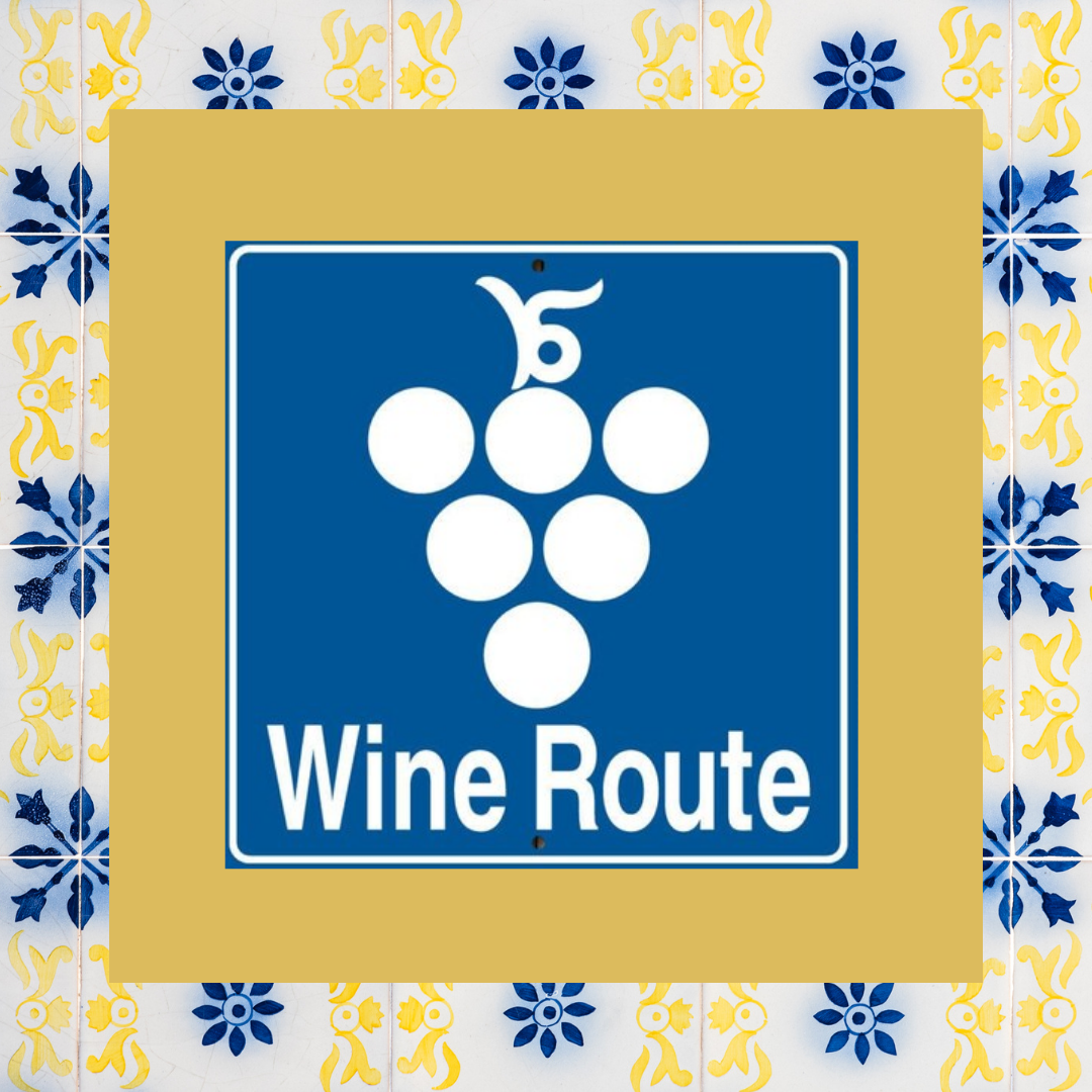 A sign with a blue background and white text and graphics that reads "Wine Route." The graphic features a cluster of white circles representing grapes and a stylized grape leaf or vine above. The sign is mounted on a wall decorated with yellow and blue floral patterned tiles.