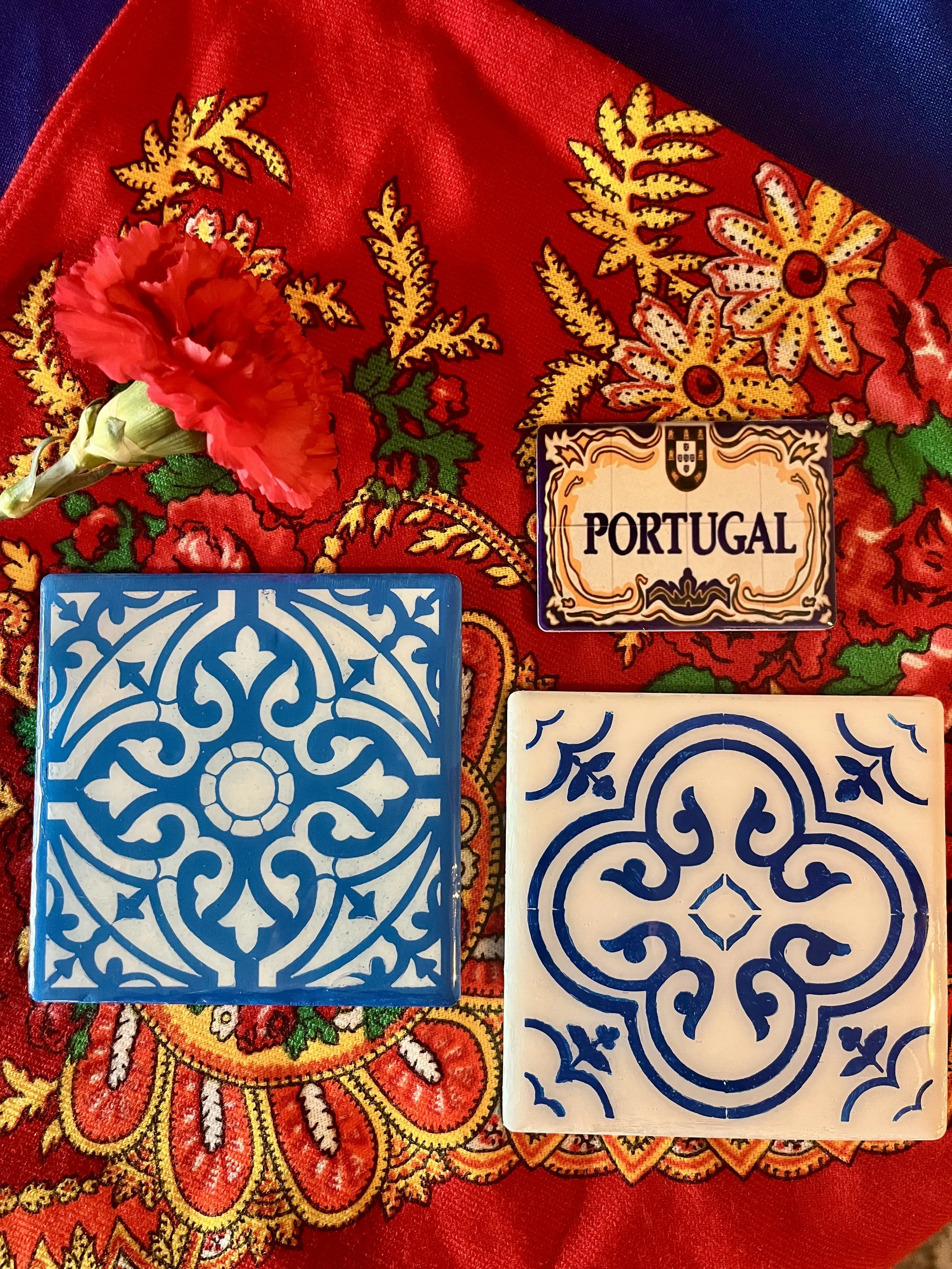 Colorful embroidered fabric with red, yellow, green, and purple patterns, a red carnation flower, and two ceramic tiles with intricate blue and white designs labeled 'Portugal' and generic patterns.