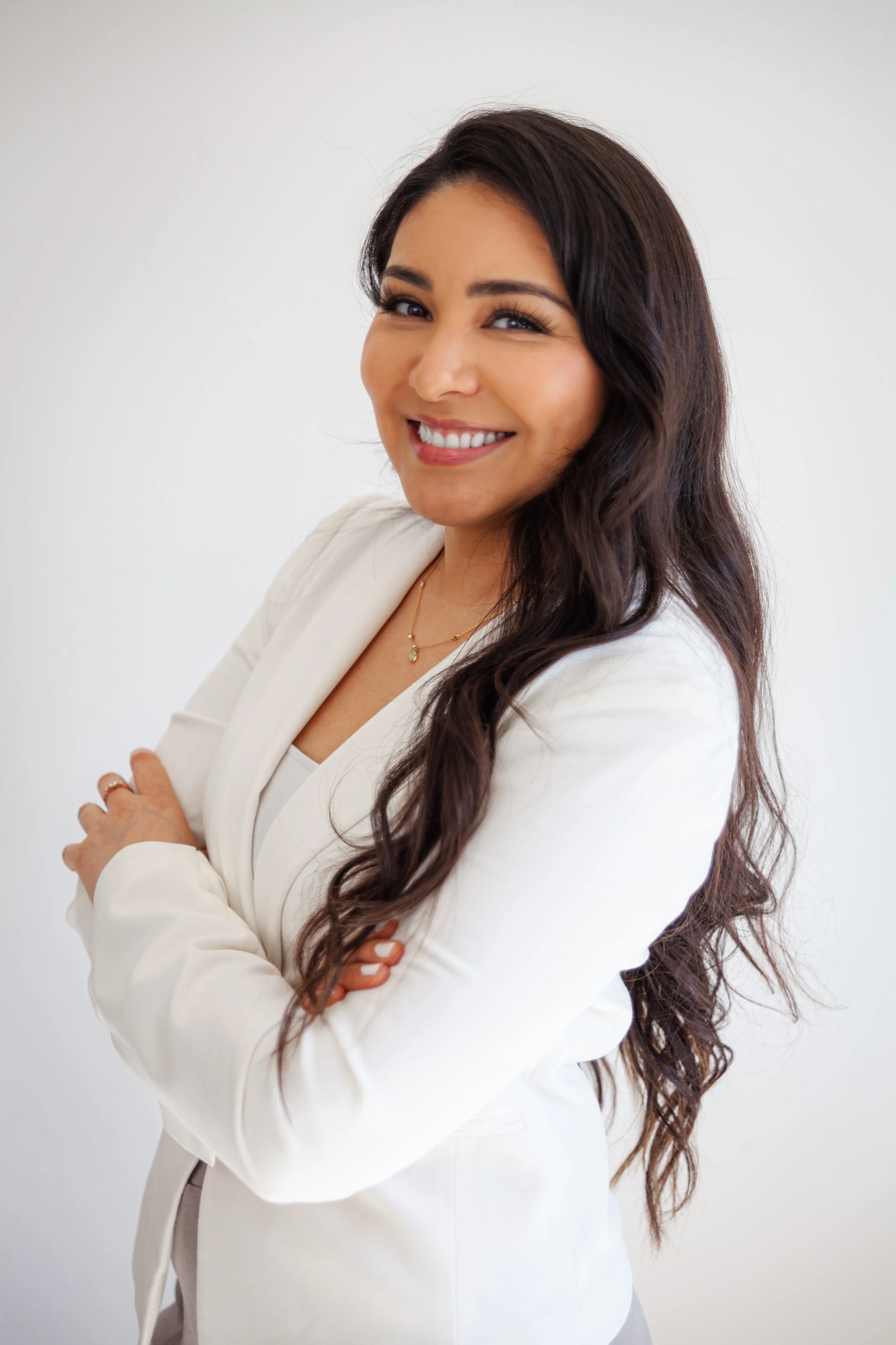 A woman with long wavy dark hair smiling and standing with arms crossed, wearing a white blazer and a delicate necklace, against a plain white background.