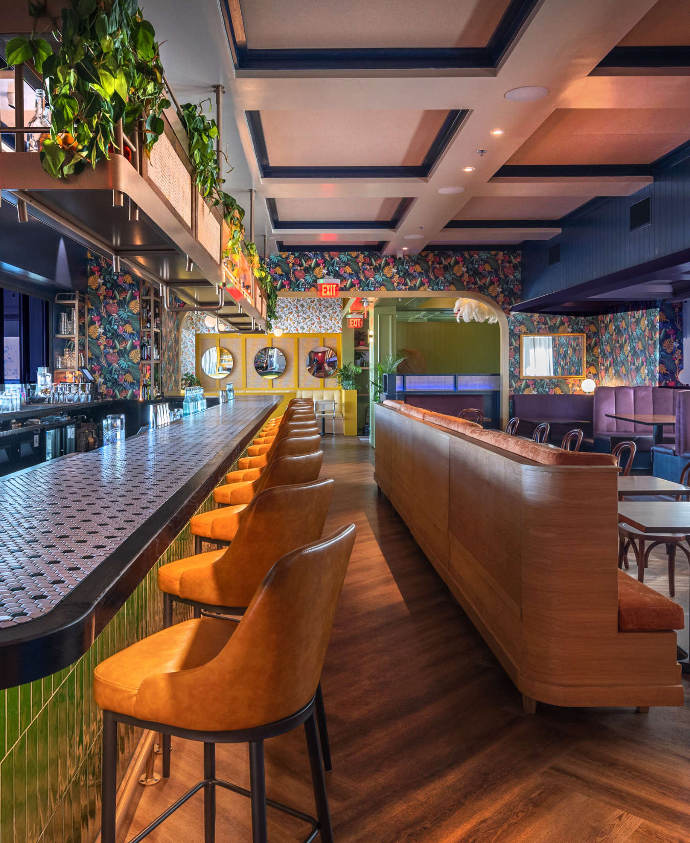 Empty bar with orange chairs along a tiled bar counter, colorful floral wallpaper, and seating area with purple and green walls in a modern restaurant.