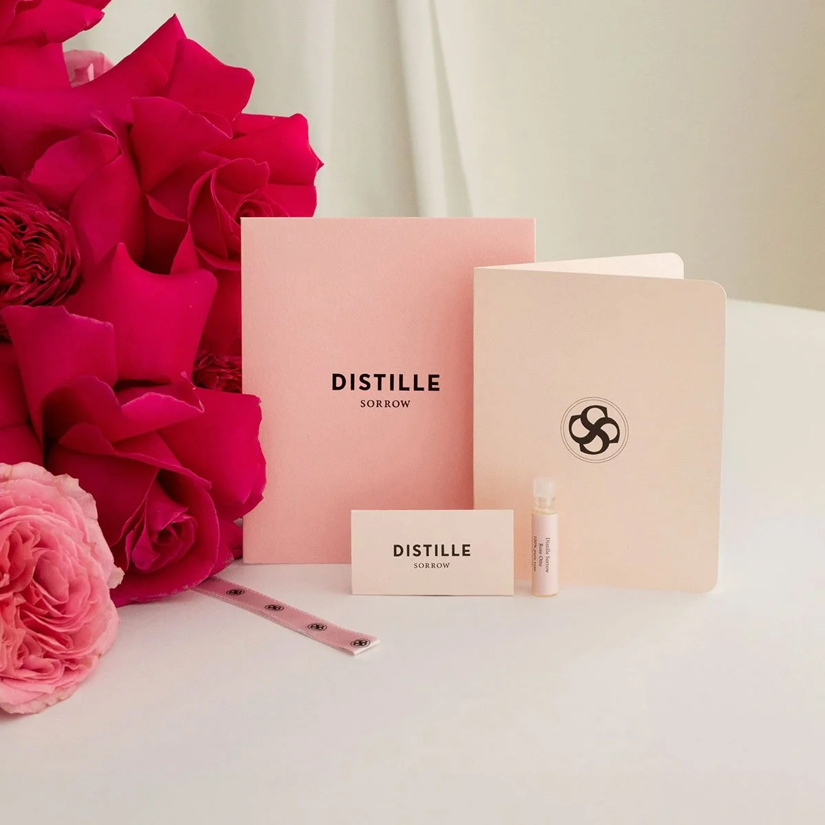 Collection of products available from Distille, including a vial of distilled rose oil, a pink greeting card, an envelope with the Distille logo, and a pink ribbon.