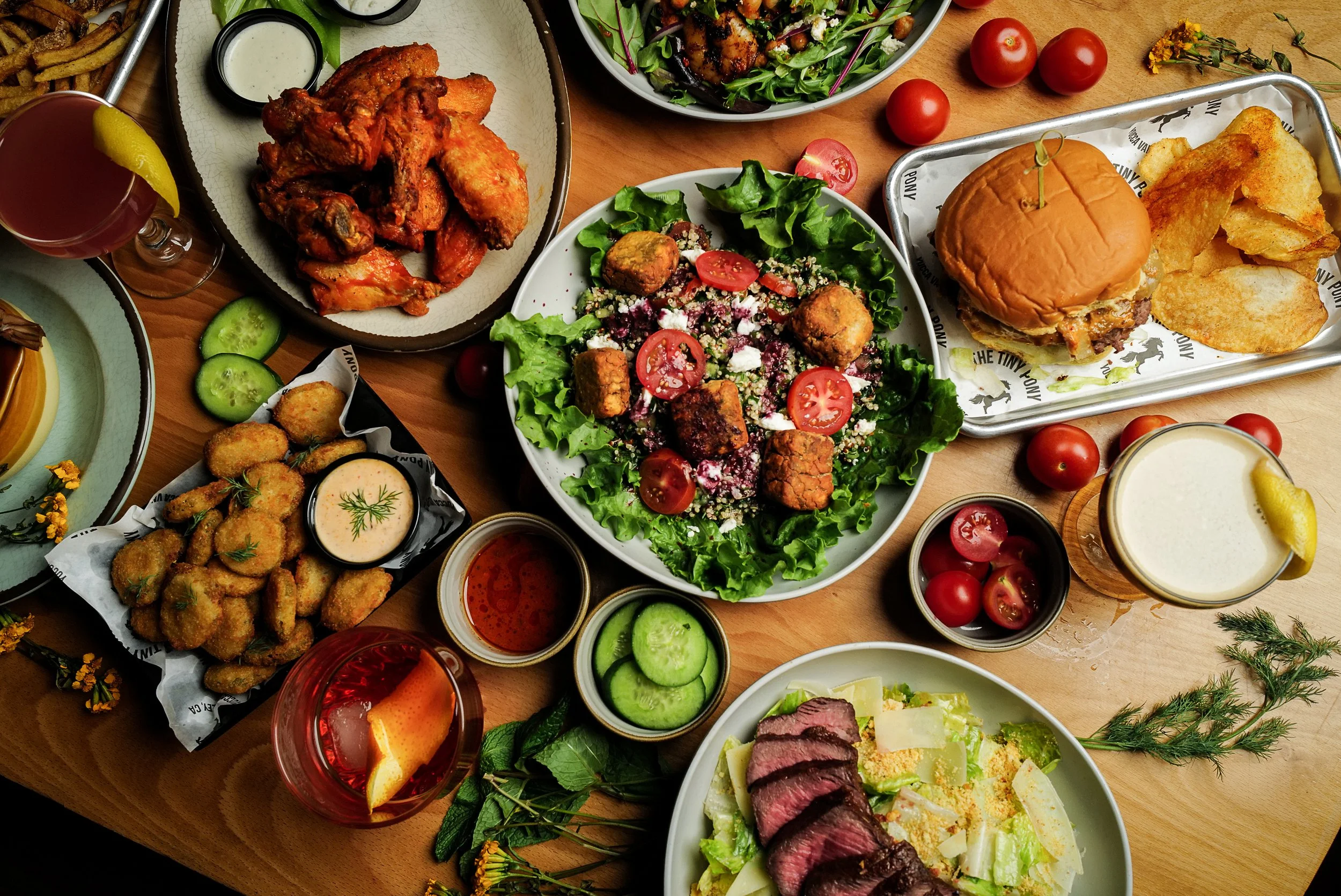 An aerial view of a salad bowl with falafel bites,  a burger with potato chips, fried pickle chips with dipping sauce, buffalo wings, and medium rare steak sliced over a caesar salad, with various garnishes, an espresso martini, and an old fashioned 