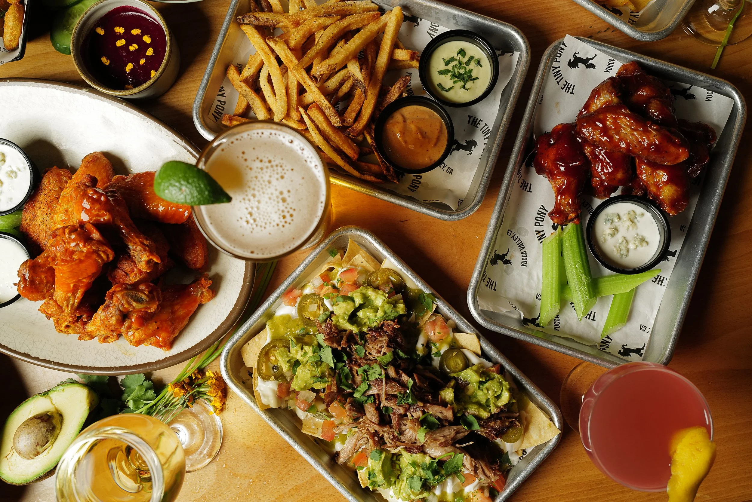 An aerial view of trays and plates featuring piled high duck confit nachos, buffalo and barbecue wings, fries with dipping sauces, wine, a pink cocktail, and a pint of beer with a lime wedge.