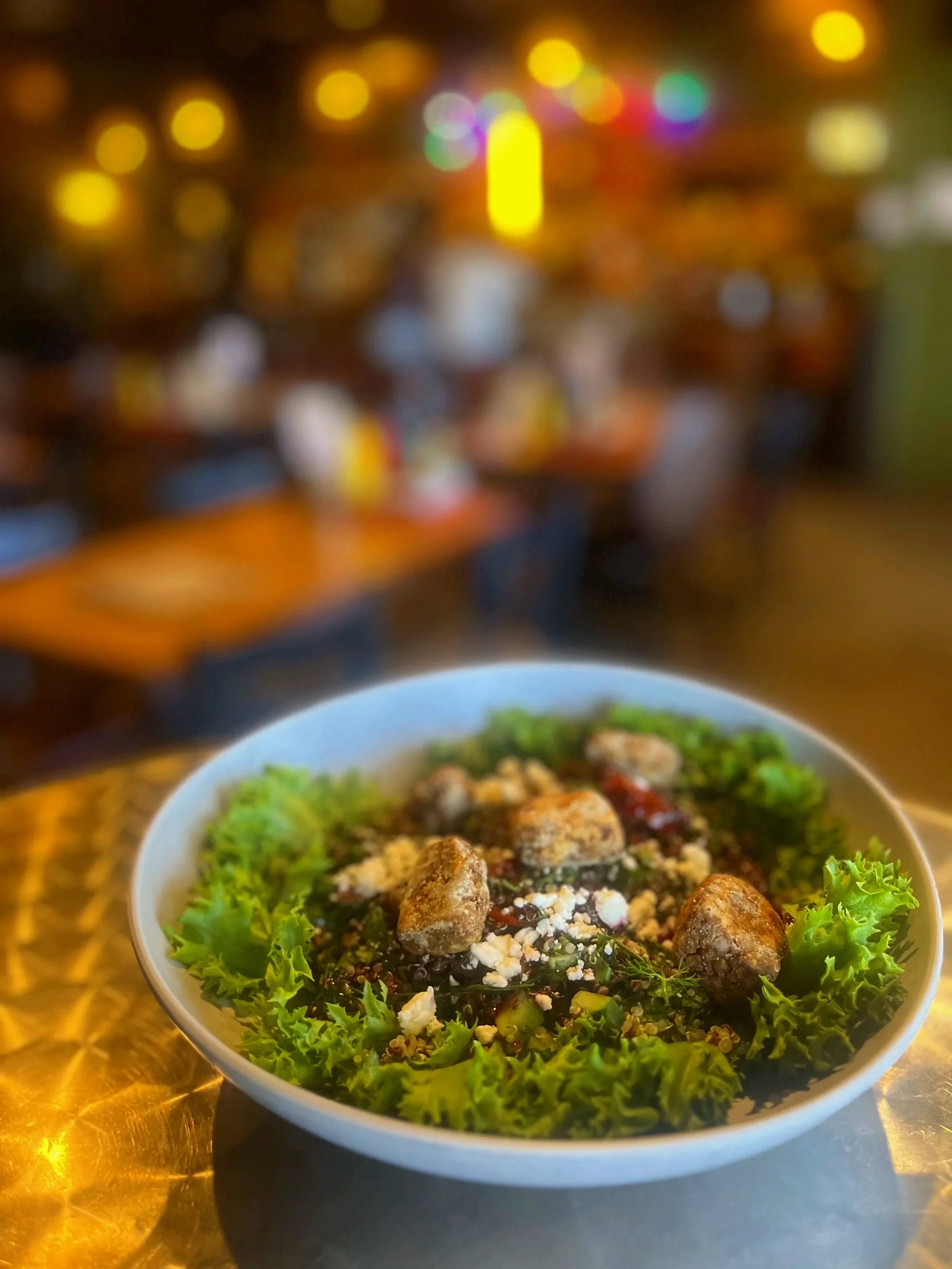 A large bowl with greens, falafel, goat cheese crumbles, and quinoa, on a holographic table with a blurry restaurant in the background.