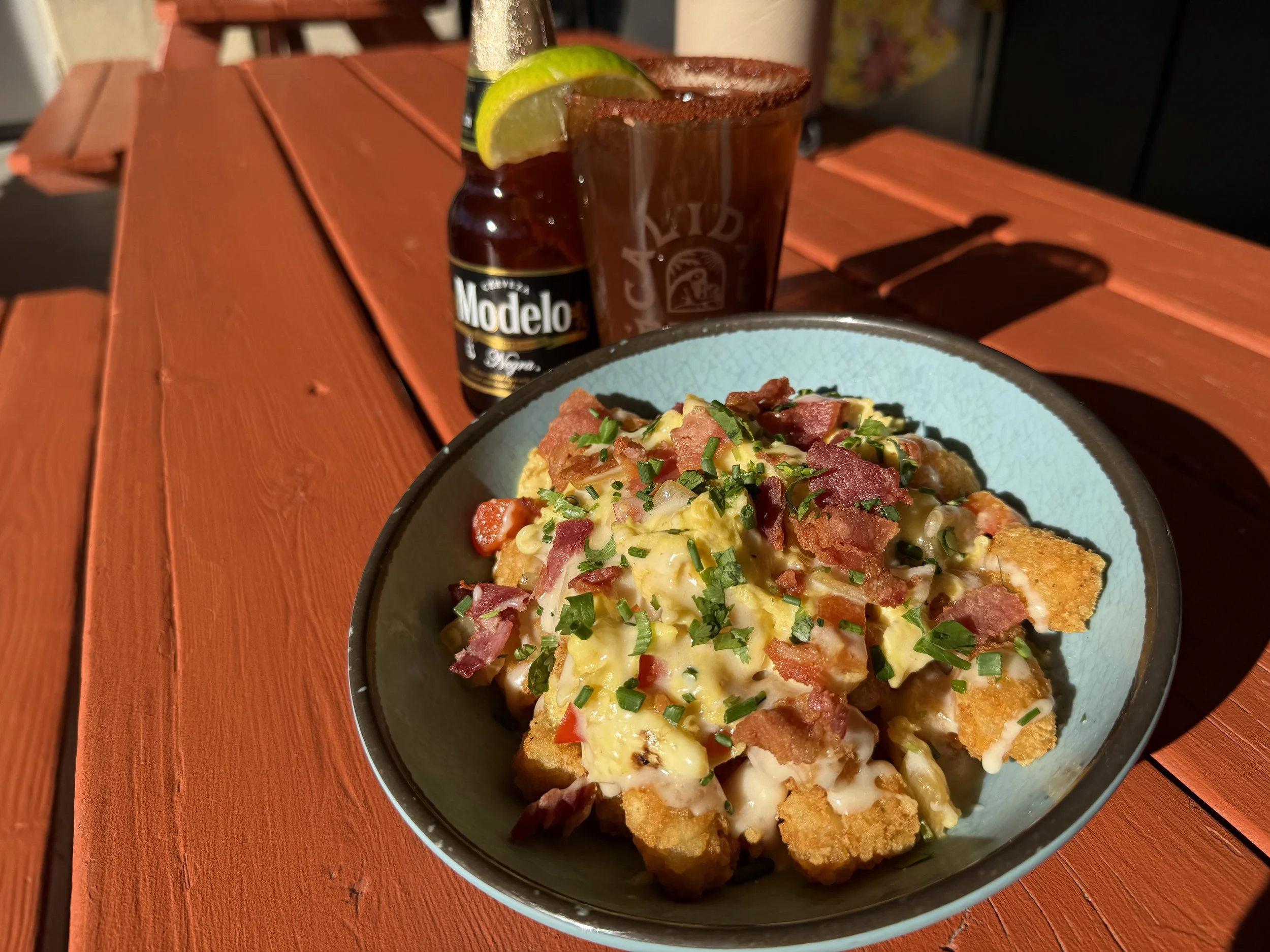 A shallow blue bowl full of melted cheesy tater tots, bacon, eggs, and chives, next to a michelada and a modelo negra beer bottle on an orange picnic table.