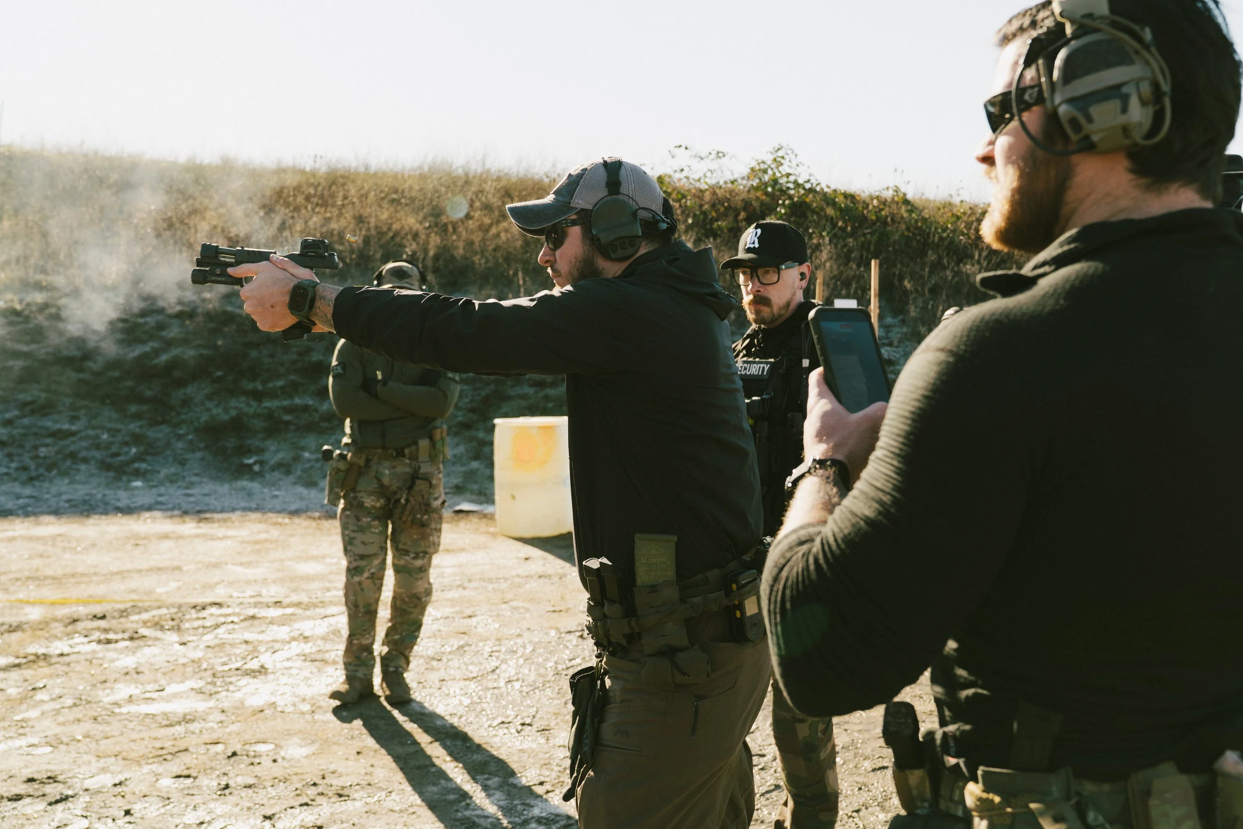 Two men wearing tactical gear and ear protection examining equipment outdoors.