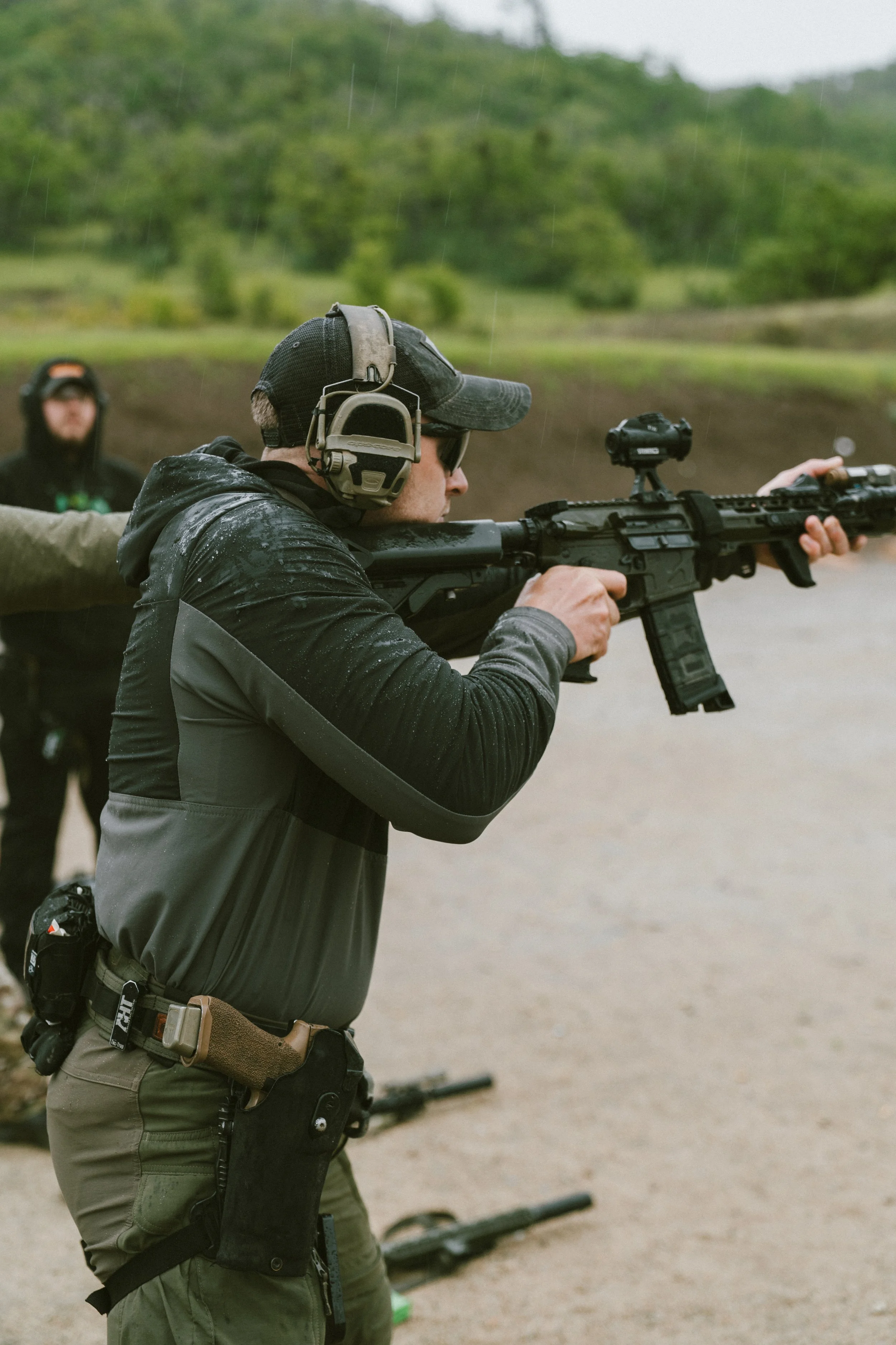 A man wearing tactical gear, black cap, sunglasses, and a black jacket at an outdoor shooting range, inspecting or marking targets on a silhouette target stand.