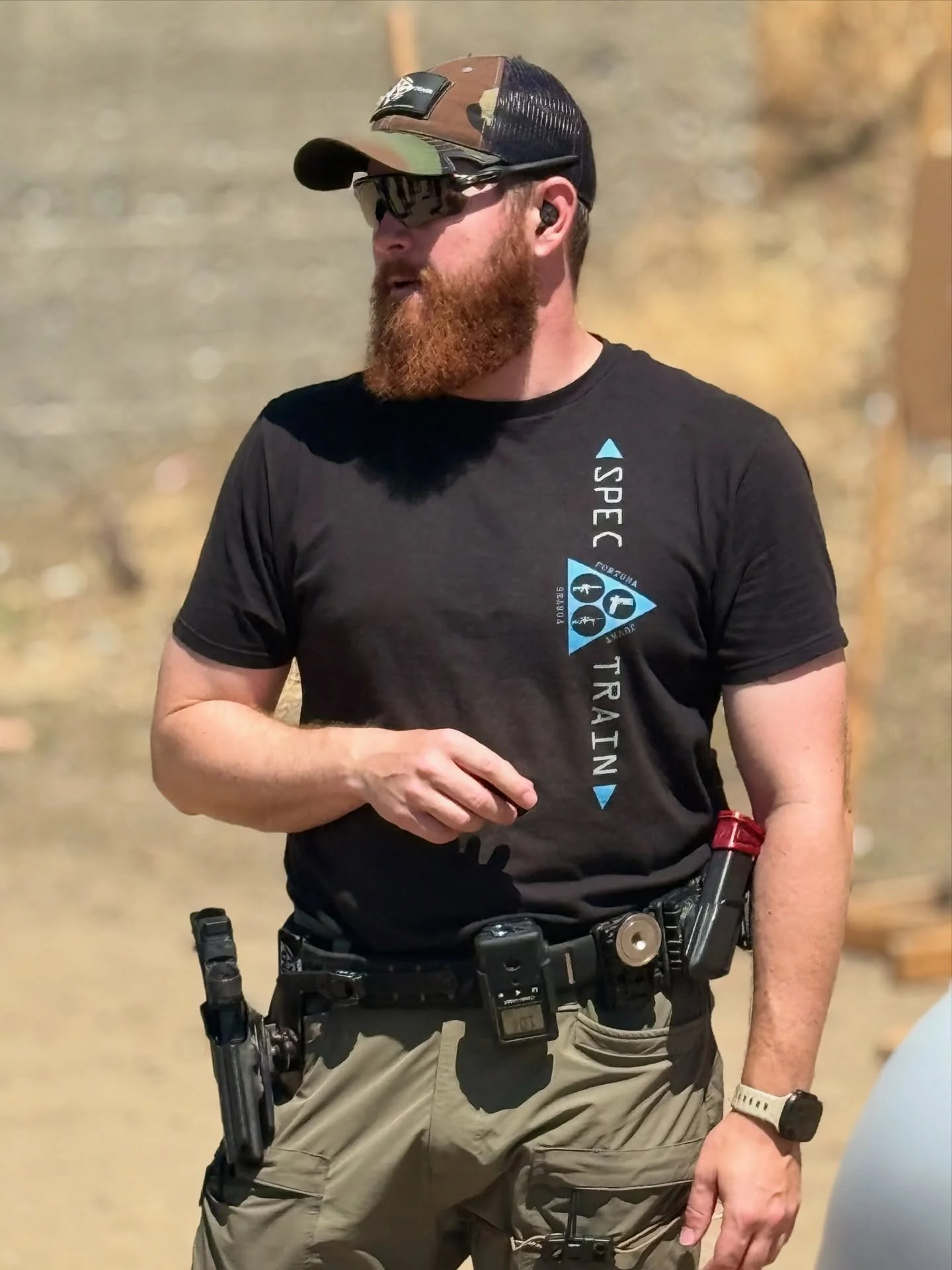 Armed security officer with a beard wearing a baseball cap, sunglasses, a black t-shirt, khaki tactical pants, and a watch, standing outdoors.