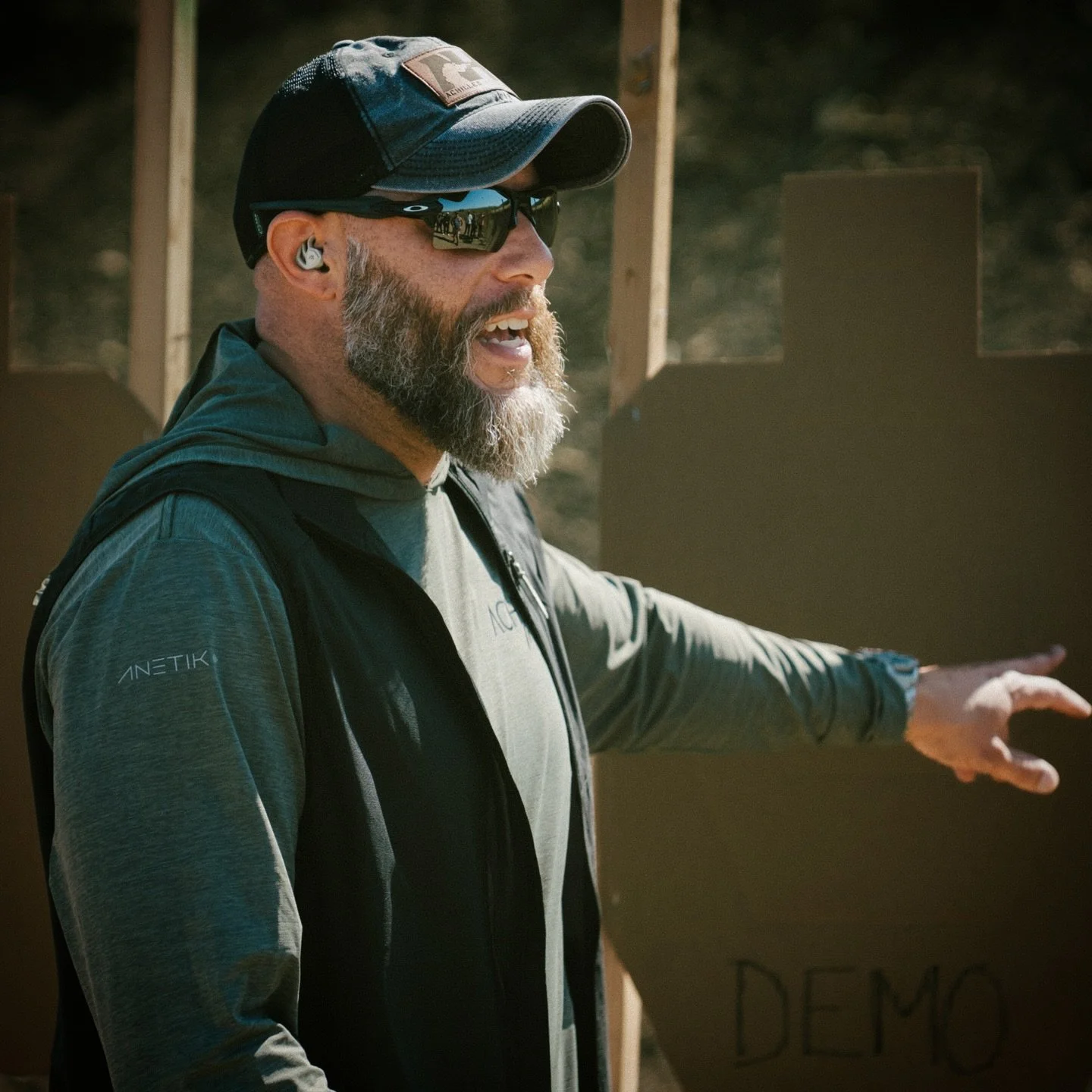 A man with a beard wearing sunglasses, a cap, and outdoor clothing is pointing or gesturing towards a sign that says 'DEMO'. He is outdoors, likely at a shooting range or outdoor training area.