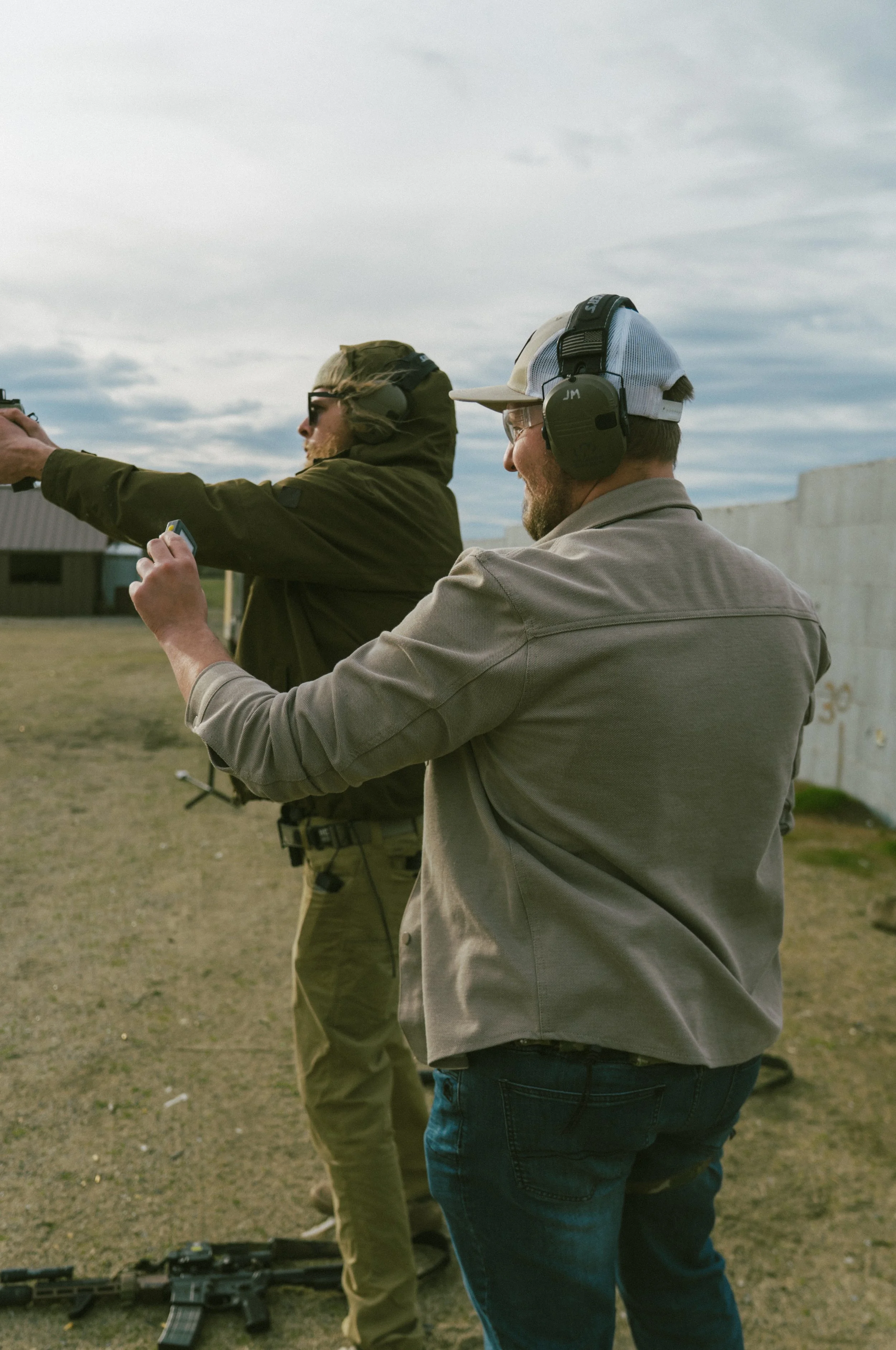 Two men wearing tactical gear and ear protection examining equipment outdoors.