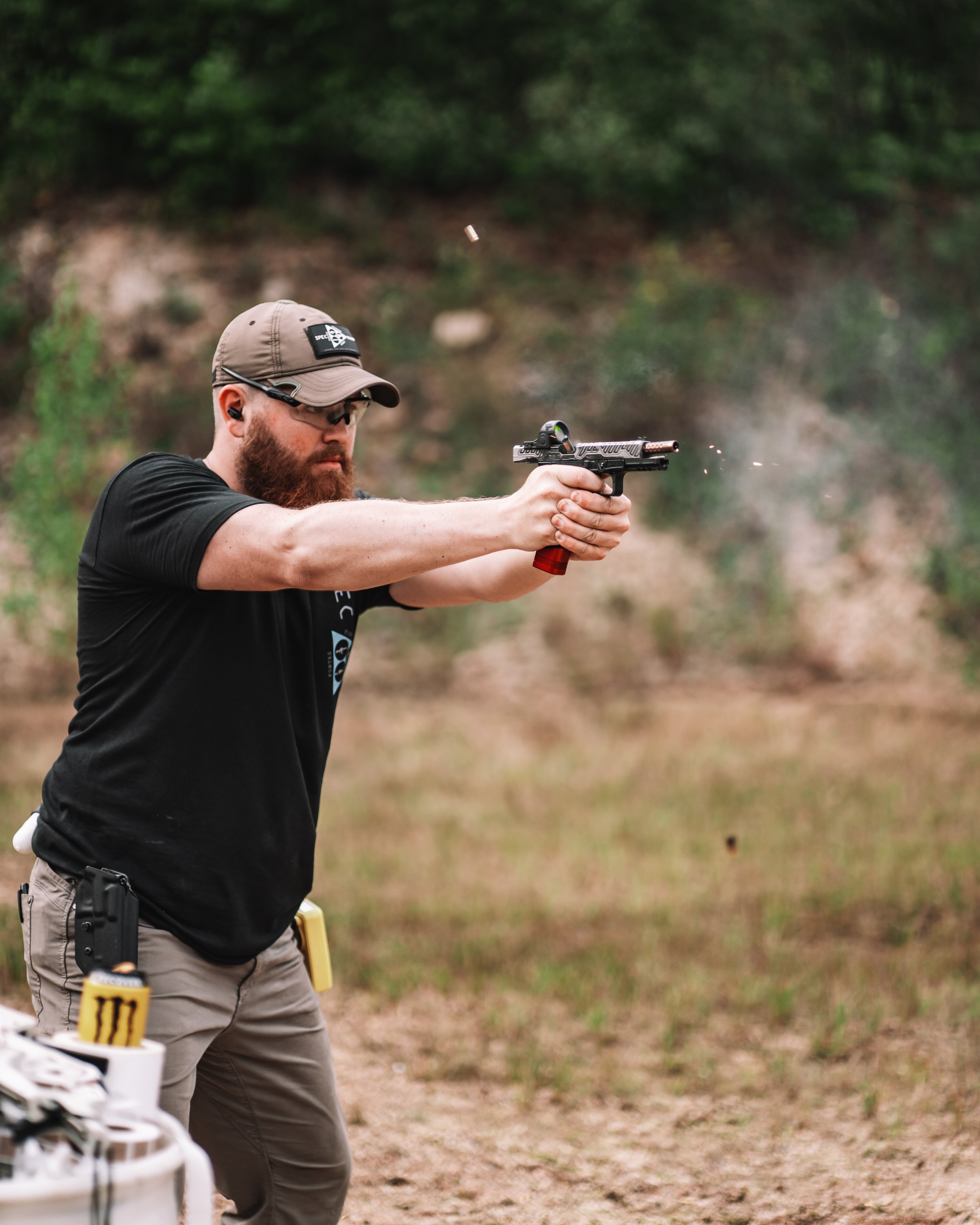 Armed security officer with a beard wearing a baseball cap, sunglasses, a black t-shirt, khaki tactical pants, and a watch, standing outdoors.
