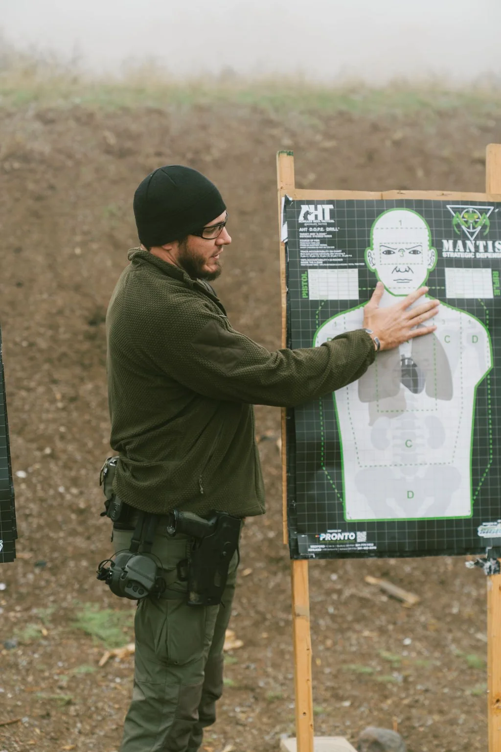 Two men wearing tactical gear and ear protection examining equipment outdoors.