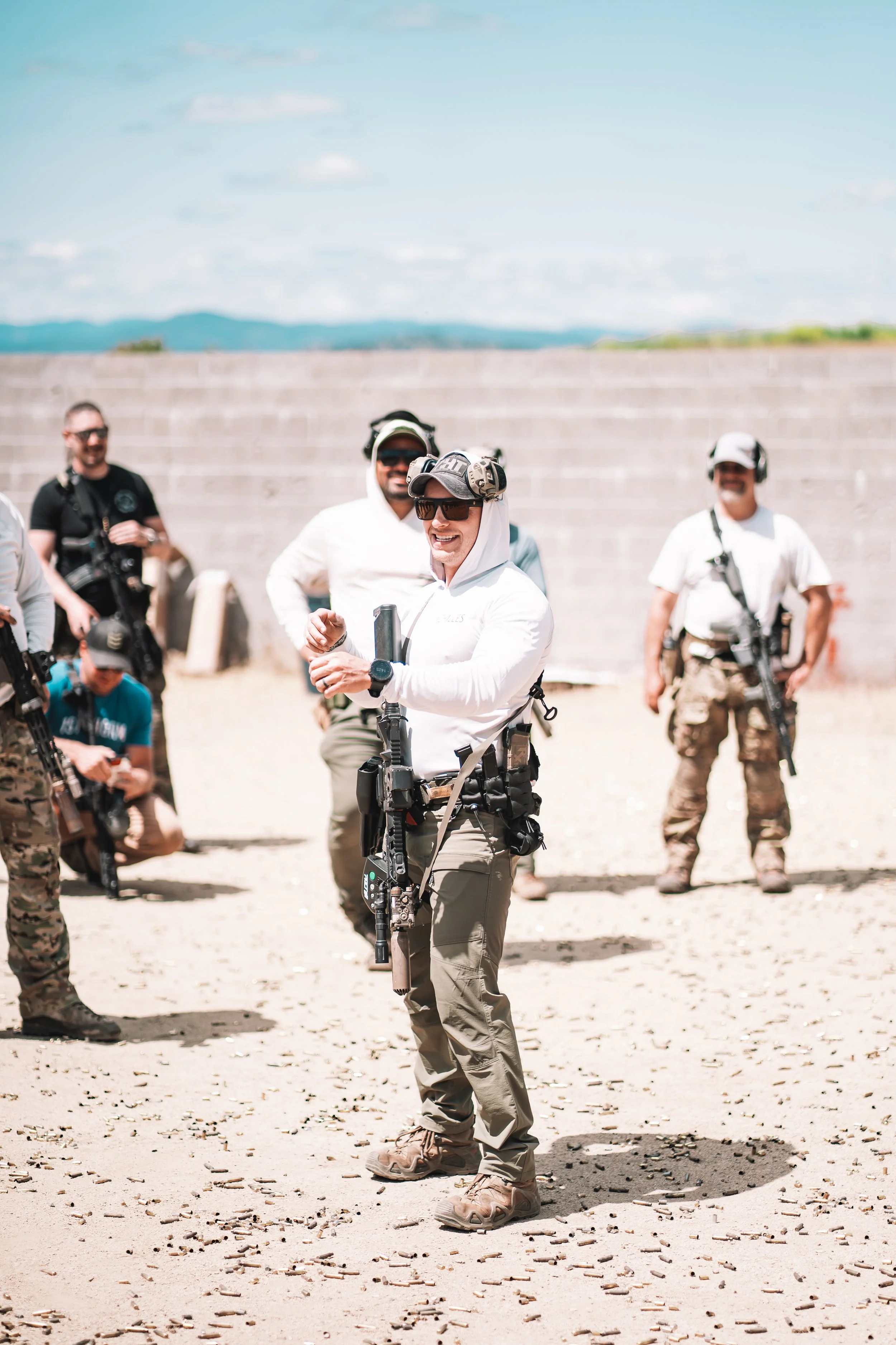 A man wearing tactical gear, black cap, sunglasses, and a black jacket at an outdoor shooting range, inspecting or marking targets on a silhouette target stand.