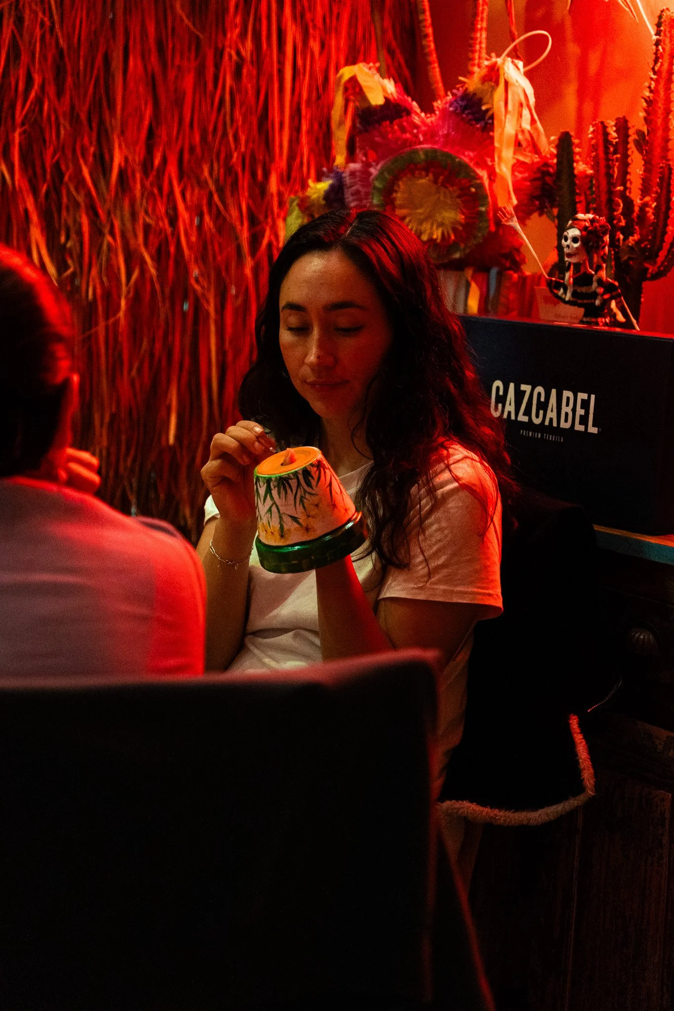A woman with long dark hair sitting in a dimly lit room with red lighting, holding a colorful ceramic cup. In the background, there are decorative elements including a skeleton figure and tall cacti, with a sign that says 'GAZCABEL'.