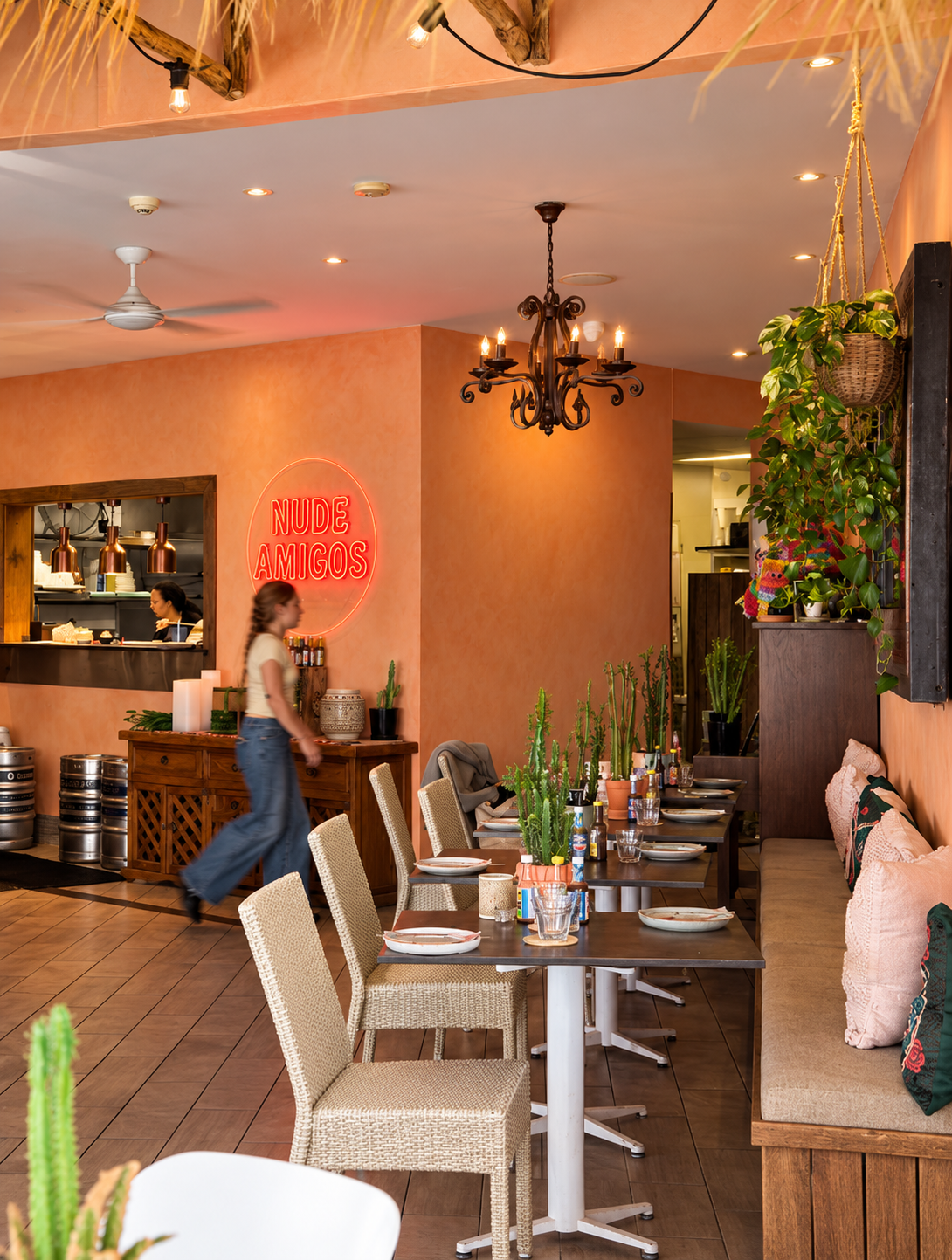 Interior of a cozy restaurant with a coral-colored wall, a neon sign that reads 'NUDE AMIGOS,' and a mix of wicker and wooden furniture. There are plants and a chandelier hanging from the ceiling, with a window opening to the kitchen where a person i