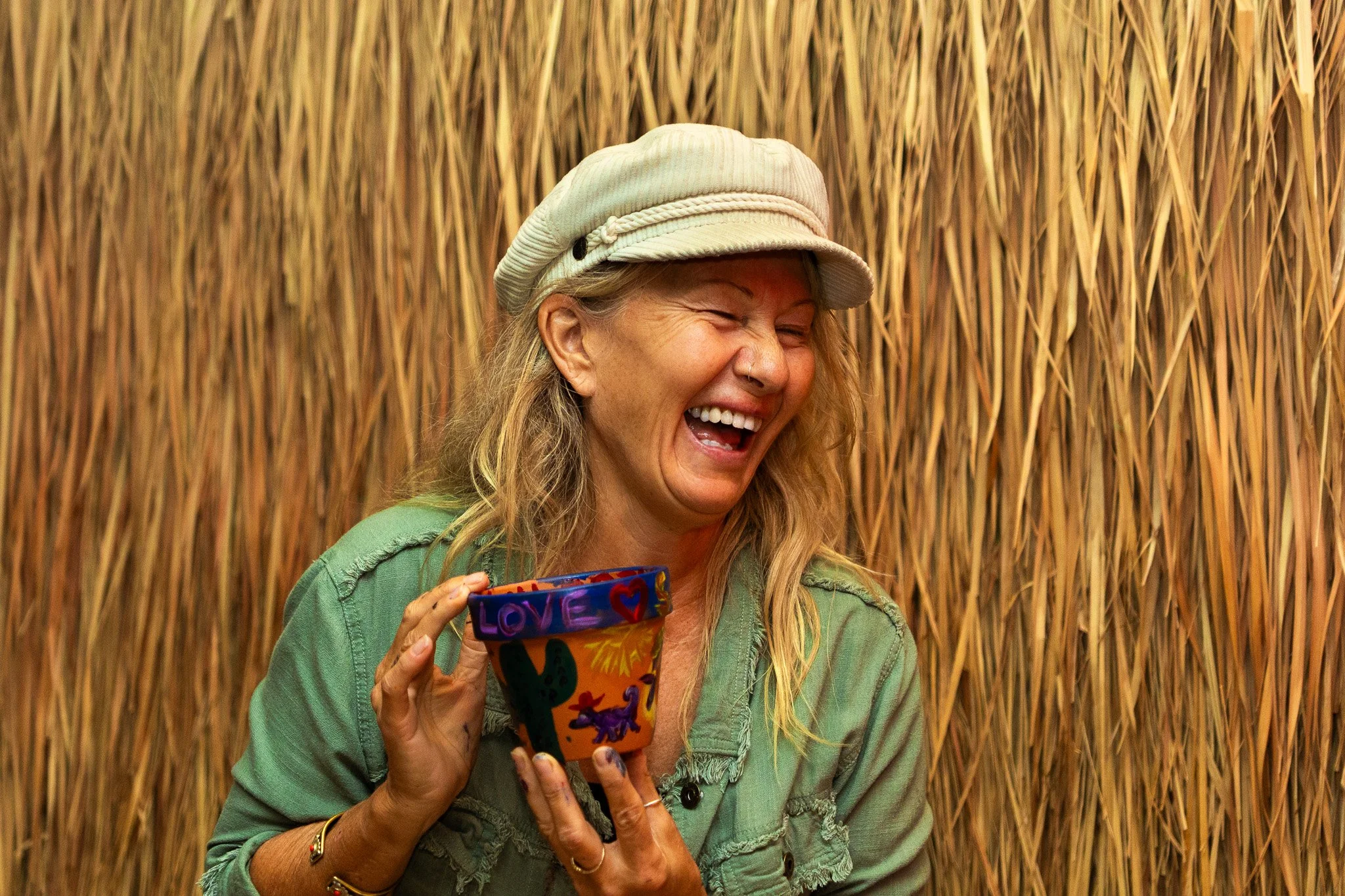 Woman with blond hair and hat laughing and holding a colorful cup with love and heart designs, standing in front of a straw or wheat background.