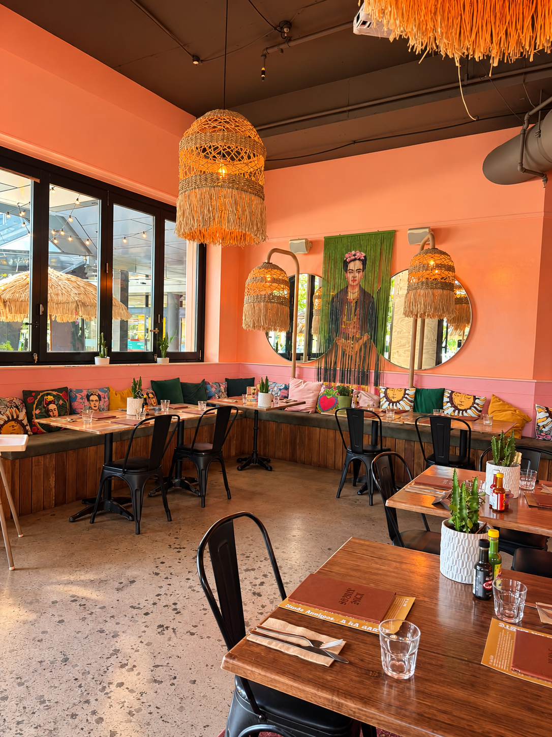 Interior of a colorful restaurant with pink walls, wooden tables, black chairs, decorative pillows featuring Frida Kahlo and other designs, hanging woven pendant lights, and a mural of Frida Kahlo on the wall with round mirrors.