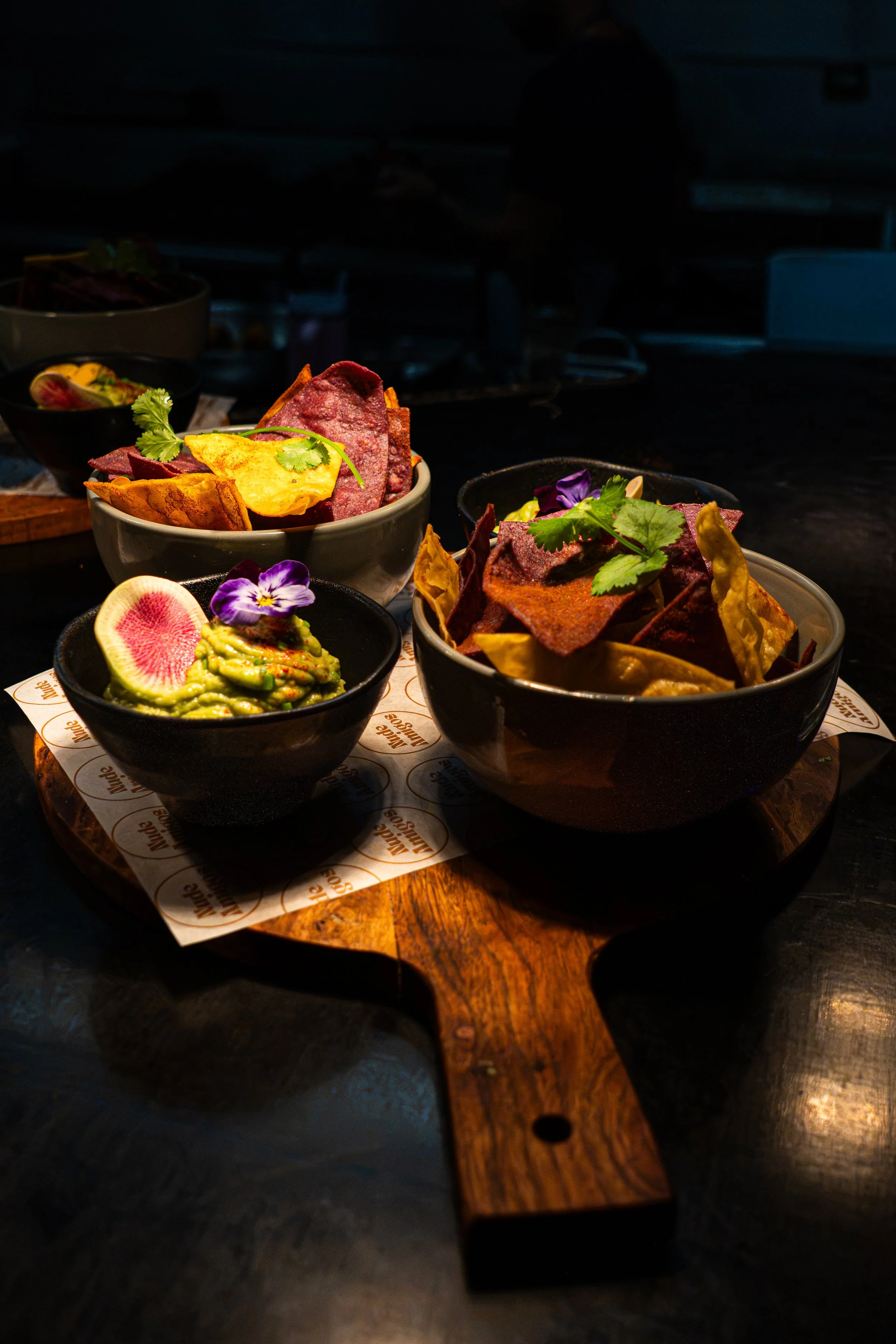 A wooden serving board with three bowls of assorted snacks, including chips, guacamole with a slice of radish and edible flowers, and other items garnished with cilantro, set on a dark table.