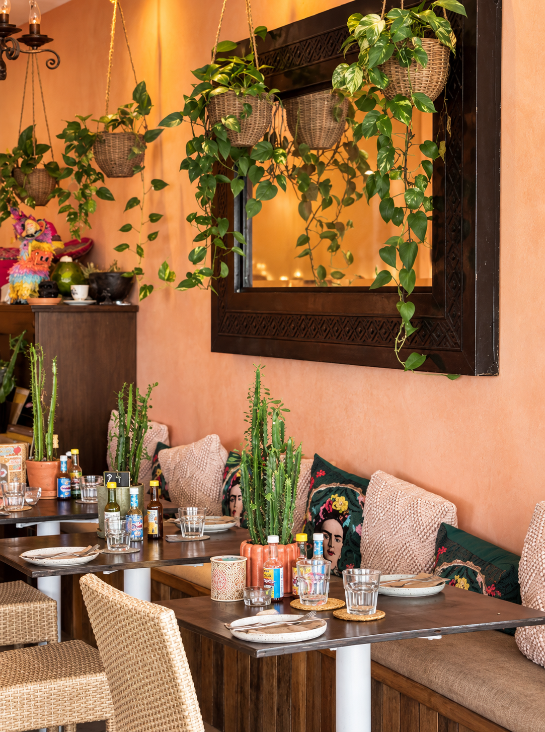 Decorative indoor restaurant table with cactus plants, colorful cushions, vintage candle holders, bottles of hot sauce, glasses, and plates, surrounded by hanging planters with trailing greenery behind a mirror on a peach-colored wall.
