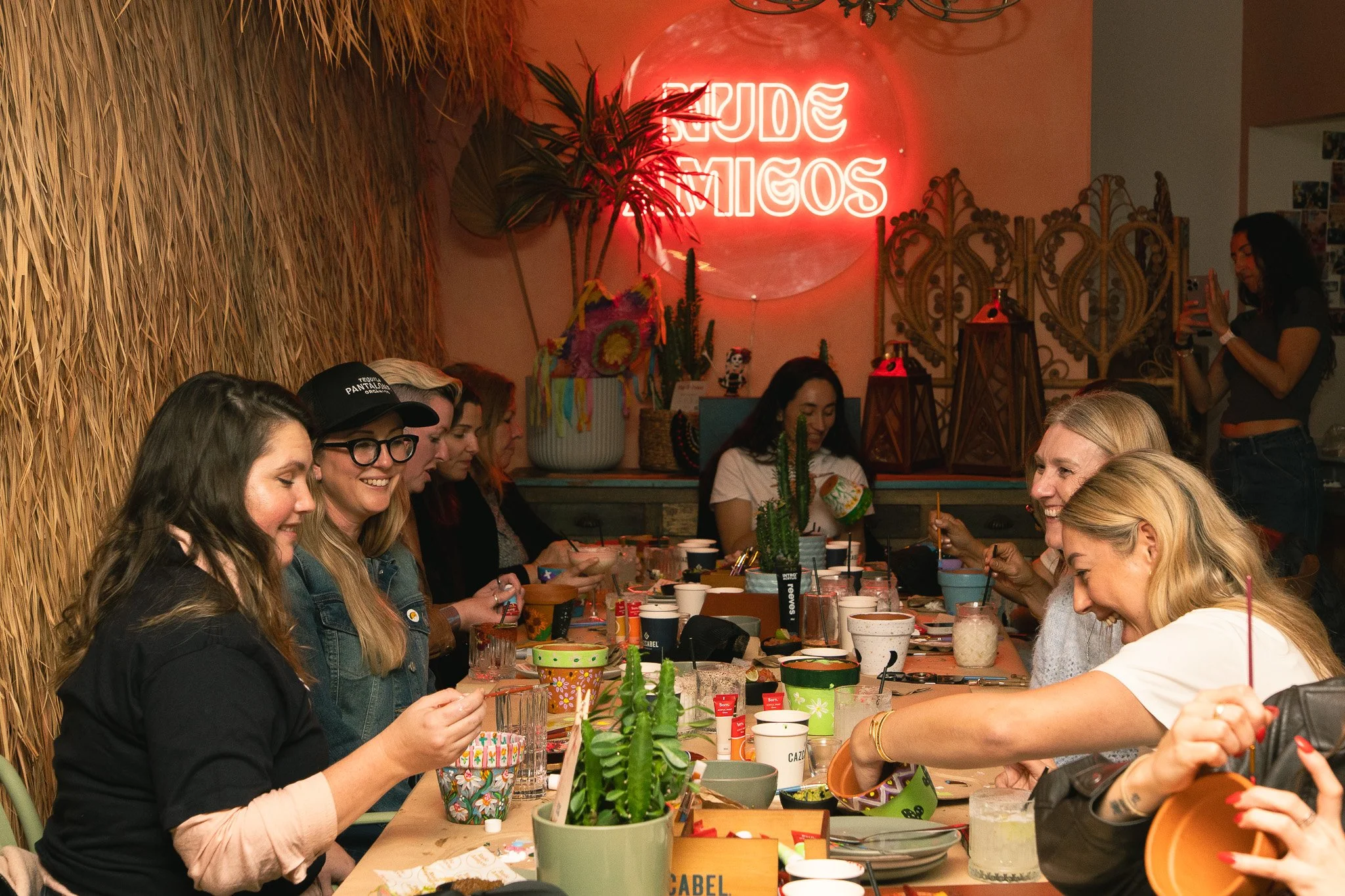 A group of women sitting at a long wooden table enjoying a gathering. The setting has a tropical theme with a wall covered in straw and a neon sign that reads 'ERASE TATTOOS'. There are various plants, decorative lanterns, and colorful cups on the ta