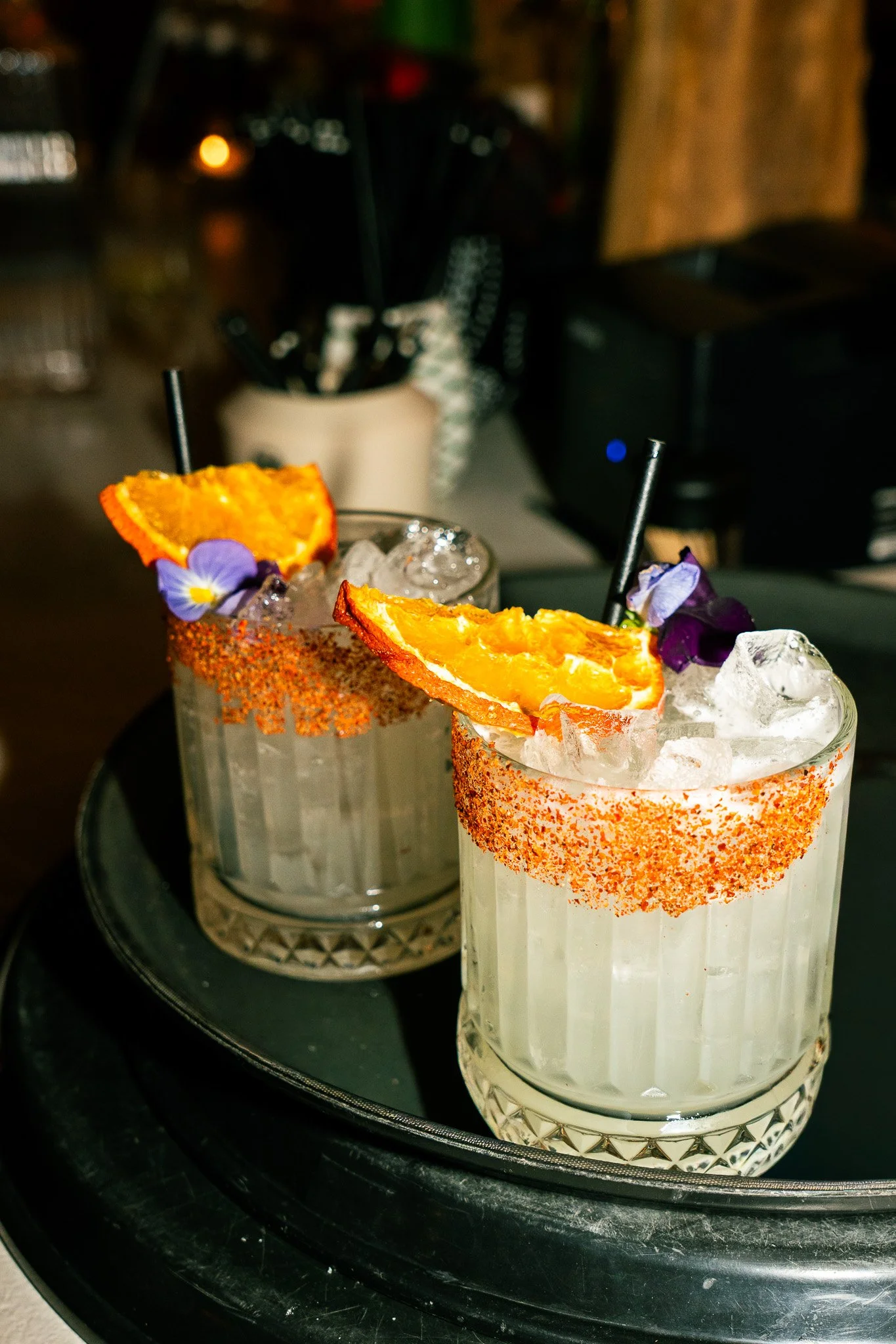 Two glasses of colorful cocktails garnished with orange slices and edible flowers, placed on a black tray.