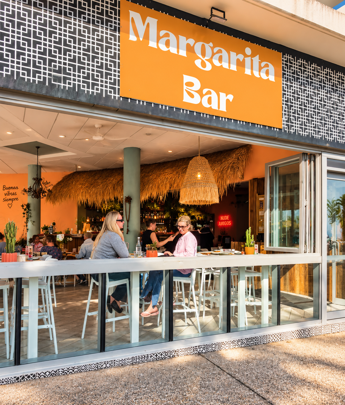 Exterior of a lively Mexican restaurant named Margarita Bar with large orange sign, open front, and outdoor seating. Two women are seated at a high table outside, chatting and smiling. Inside, there are people at tables, a bar, and tropical decor inc