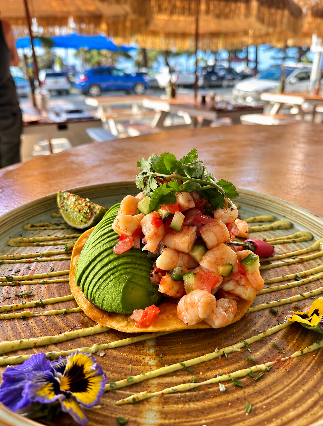 Close-up of seafood ceviche topped with cilantro, avocado slices, and lime wedge, served on a decorative plate with green sauce lines and edible flowers at a wooden table in an outdoor dining area with parked cars and blue sky in the background.