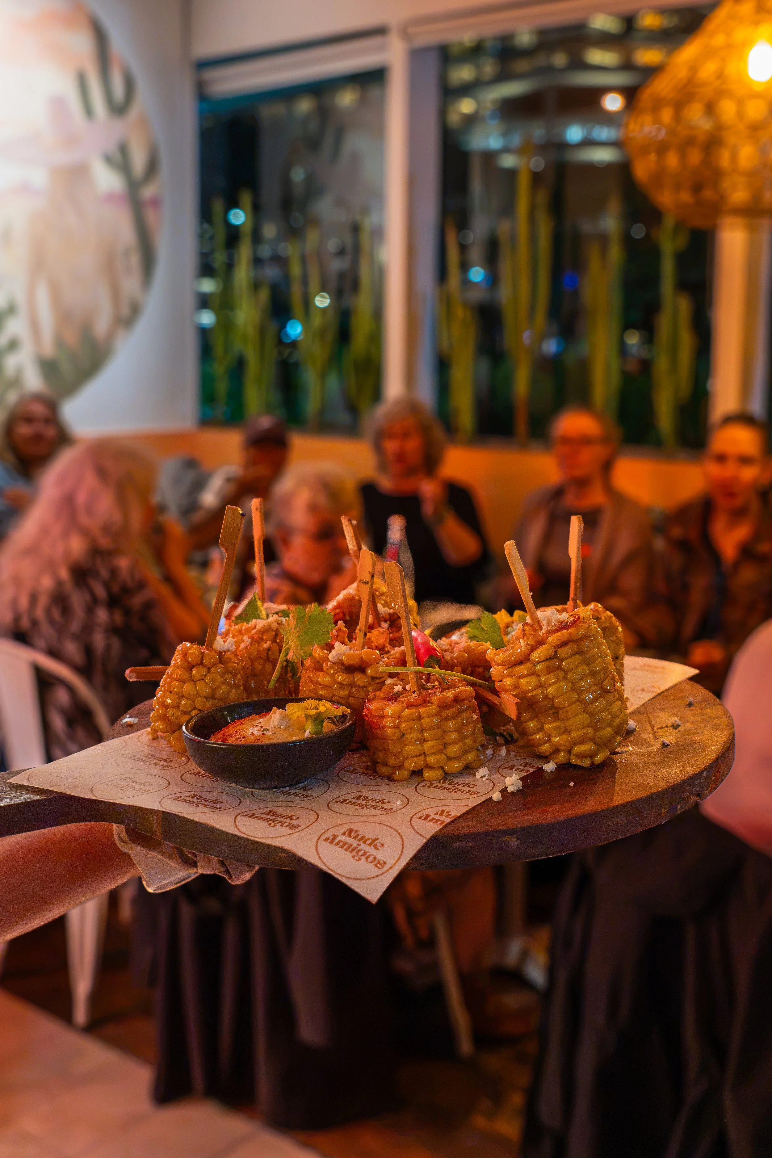 Plate of skewered corn on the cob with sauce in a small black bowl, garnished with cilantro, served at a restaurant with people in the background.