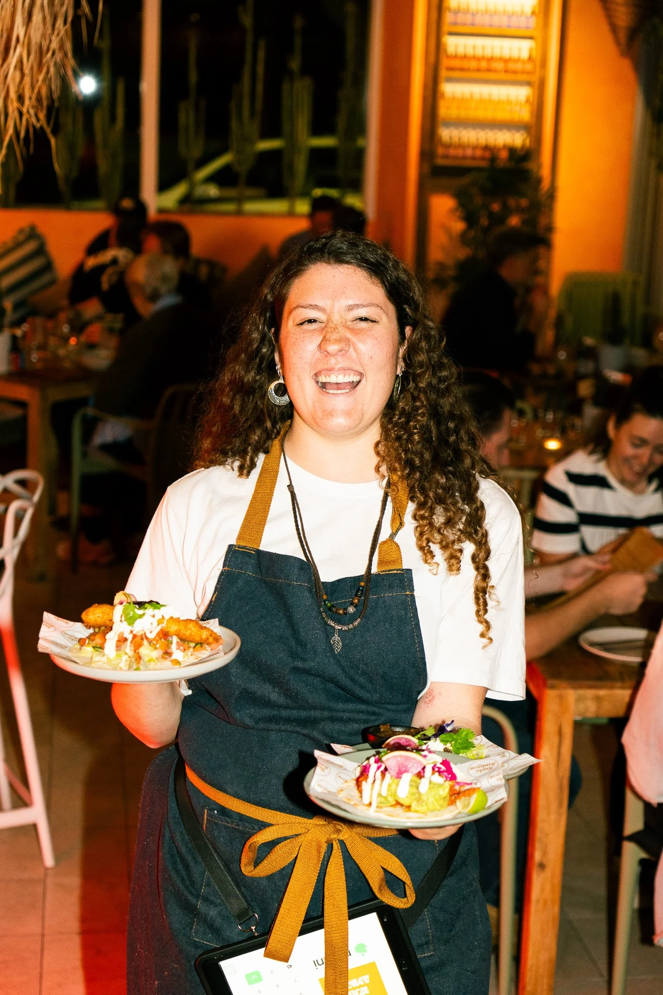 A woman with curly hair and earrings wearing a white shirt and a black apron with a yellow ribbon, holding two plates of food, smiling at the camera inside a restaurant with people dining in the background.