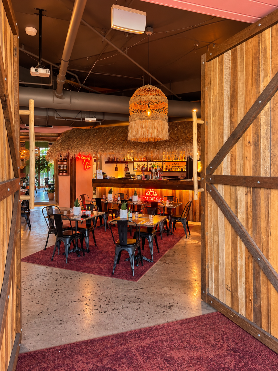 View of a cozy tropical-themed restaurant or bar with a thatched roof structure over a bar counter. Black metal chairs surround wooden tables with small potted plants. Warm lighting includes a large woven pendant lamp hanging from the ceiling. Wooden