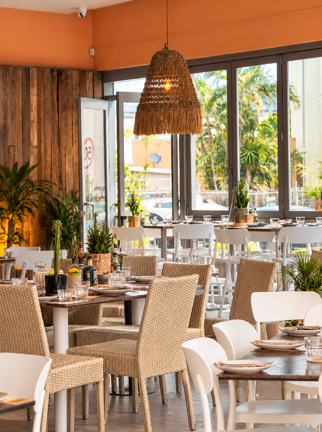 Interior of a bright restaurant with wicker and white chairs, wooden tables set with plates and glasses, potted plants, and a large woven hanging lamp.