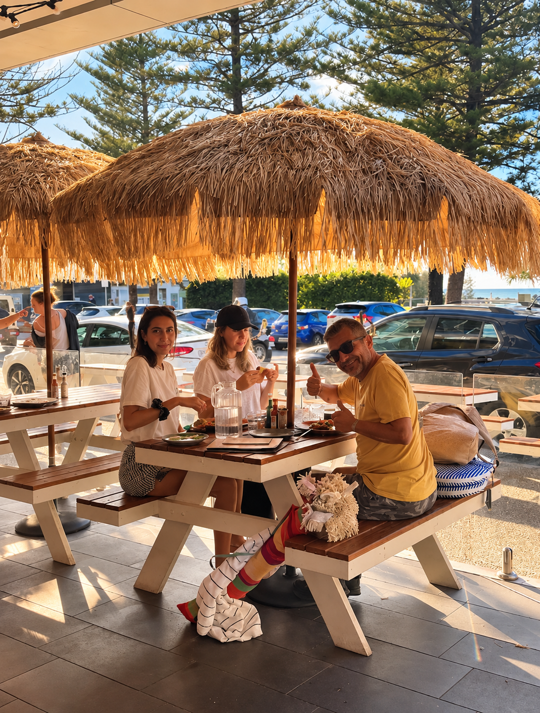 Three people sitting at a picnic table under a thatched umbrella, enjoying a meal outdoors near a parking lot, with trees and cars in the background.