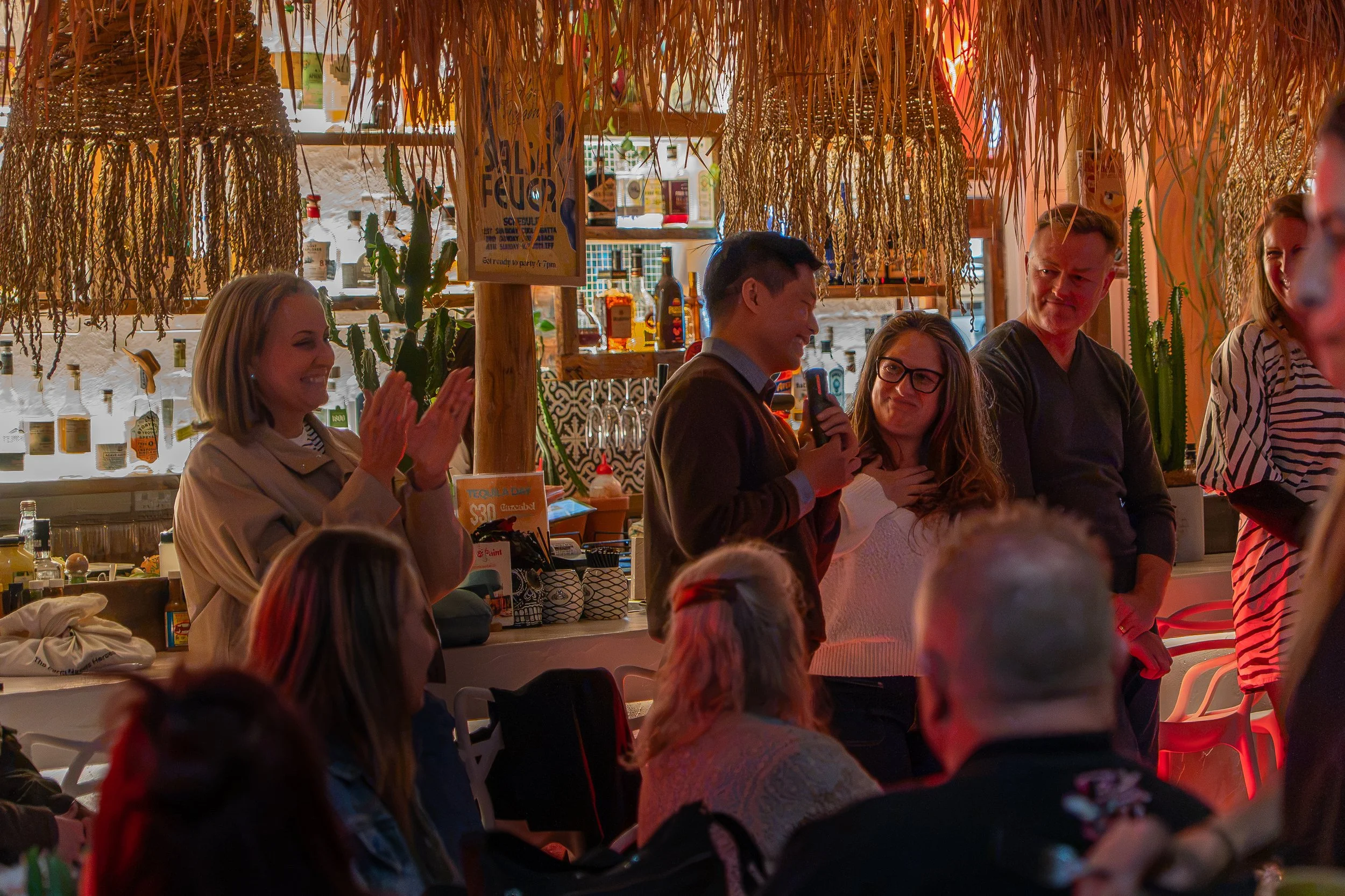 People gathered inside a restaurant or bar, some clapping and others smiling, with hanging decorations and bottles of alcohol on shelves in the background.