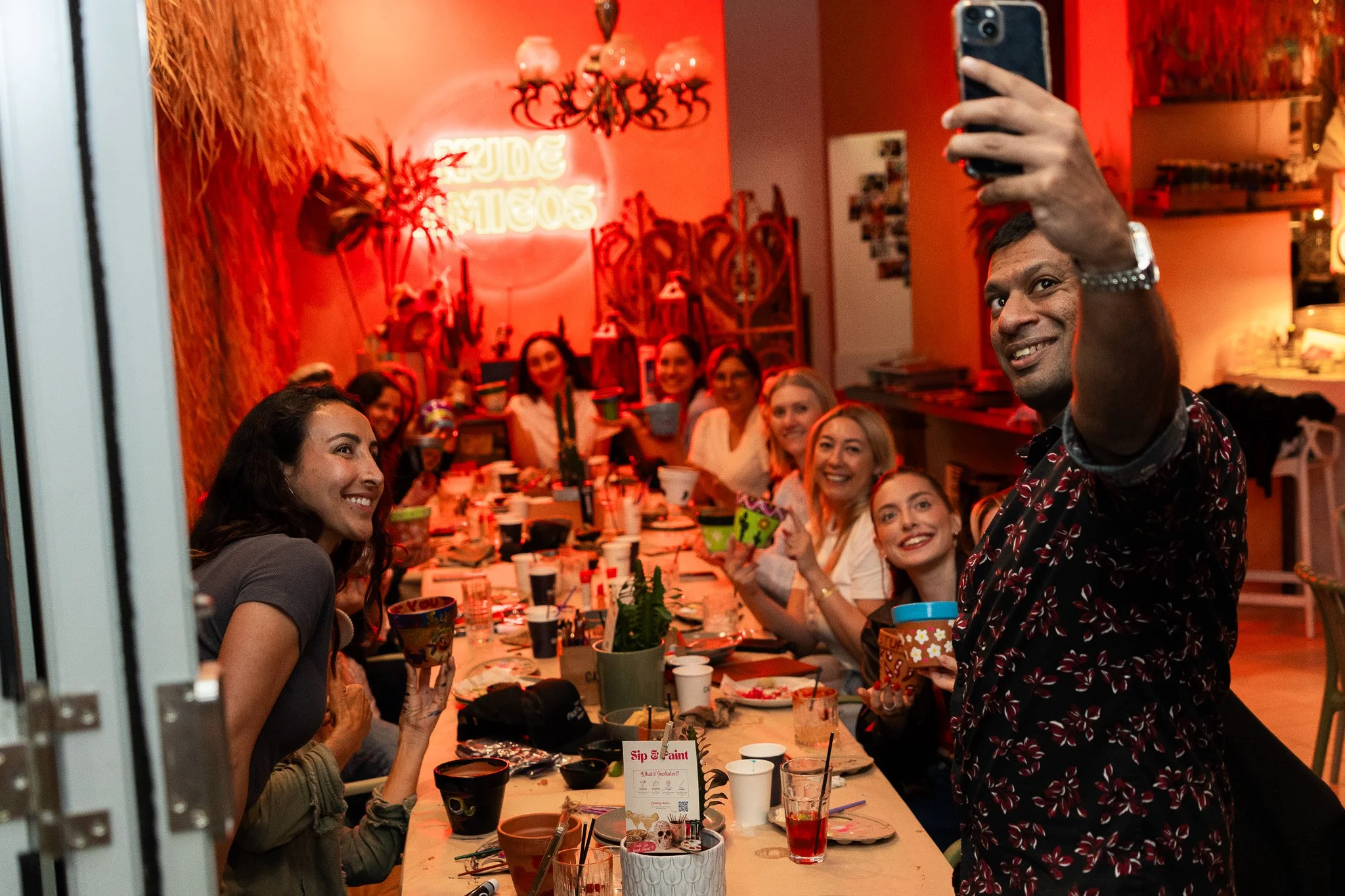 Group of people celebrating at a party, with some holding drinks and smiling. A man is taking a selfie in the foreground. The room has red lighting and decorations, with a neon sign that says 'Soul Sisters' in the background.
