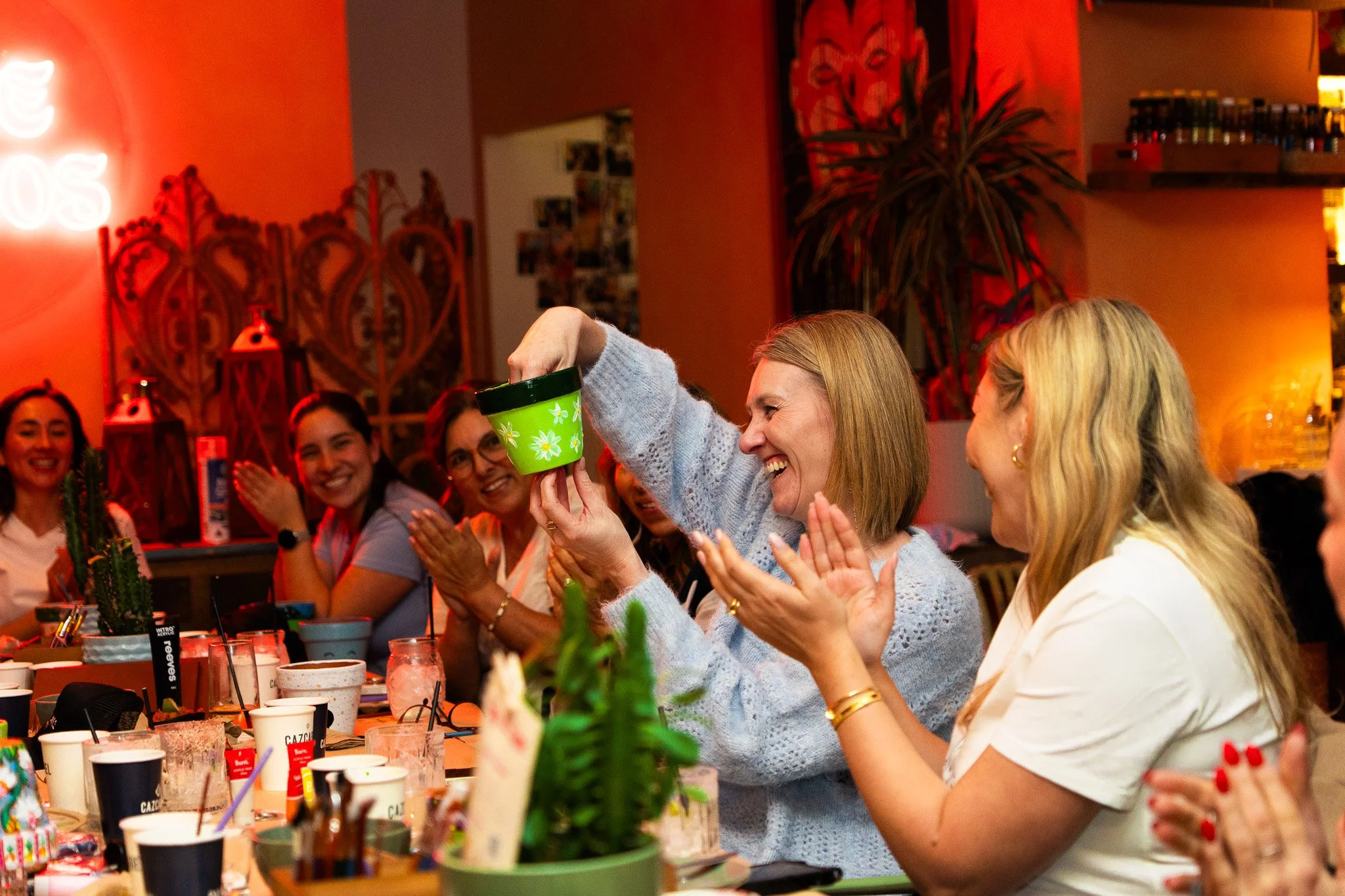 A group of women sitting at a table, smiling, with one woman raising a green cup. The setting appears festive, with colorful decorations and various drinks on the table.