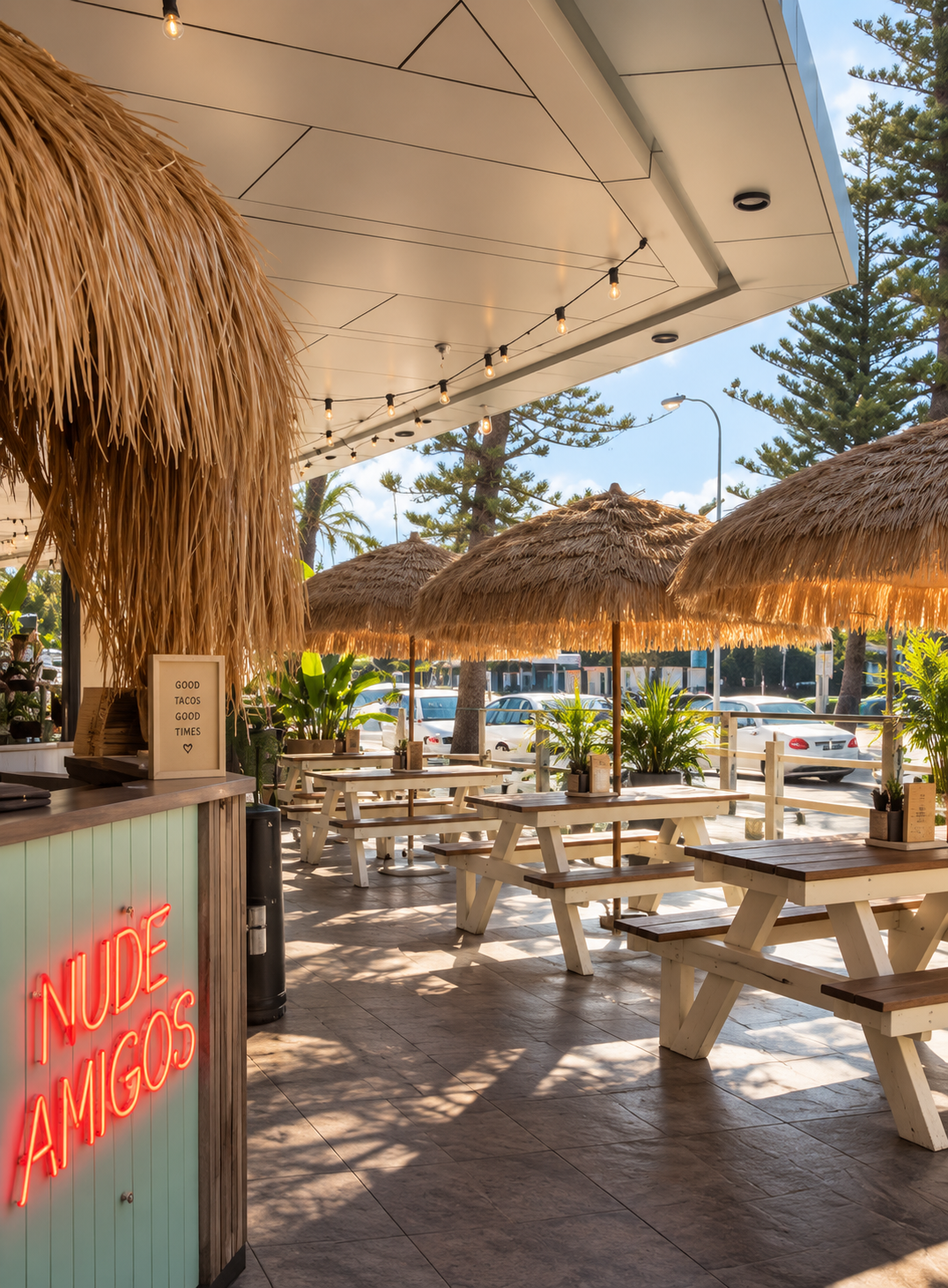 Outdoor seating area of a restaurant with wooden picnic tables, thatched umbrellas, and tropical plants, bathed in warm sunlight with string lights hanging from the ceiling.
