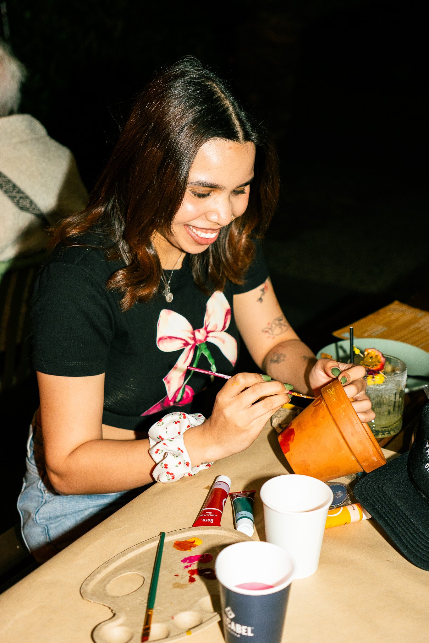 A young woman with shoulder-length brown hair, smiling while painting a ceramic pot at a table set for arts and crafts. She wears a black T-shirt with a flower design.