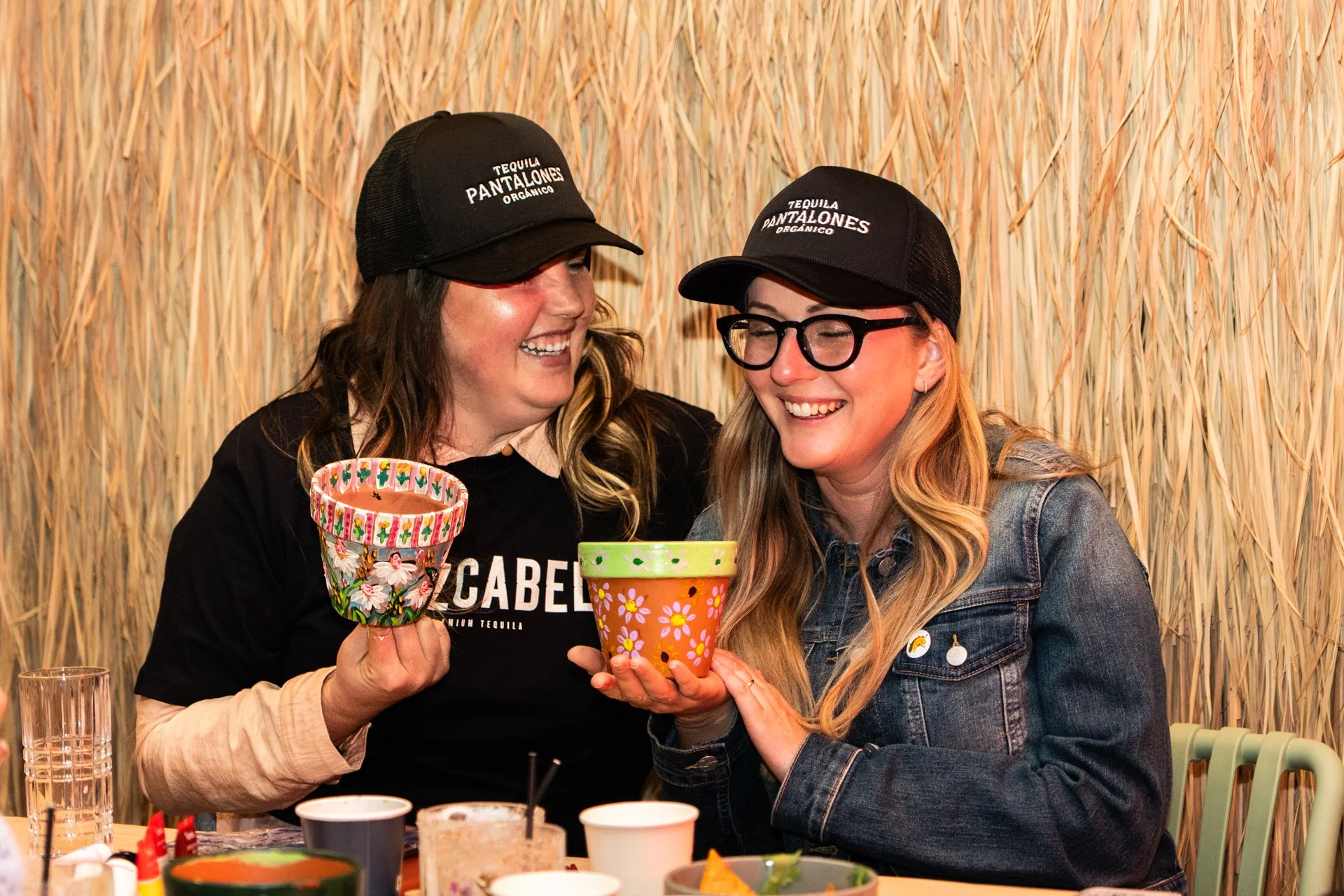 Two women smiling and holding colorful decorated cups, wearing black caps with white text, sitting at a table with various drinks and food. They are in front of a straw-like textured wall.