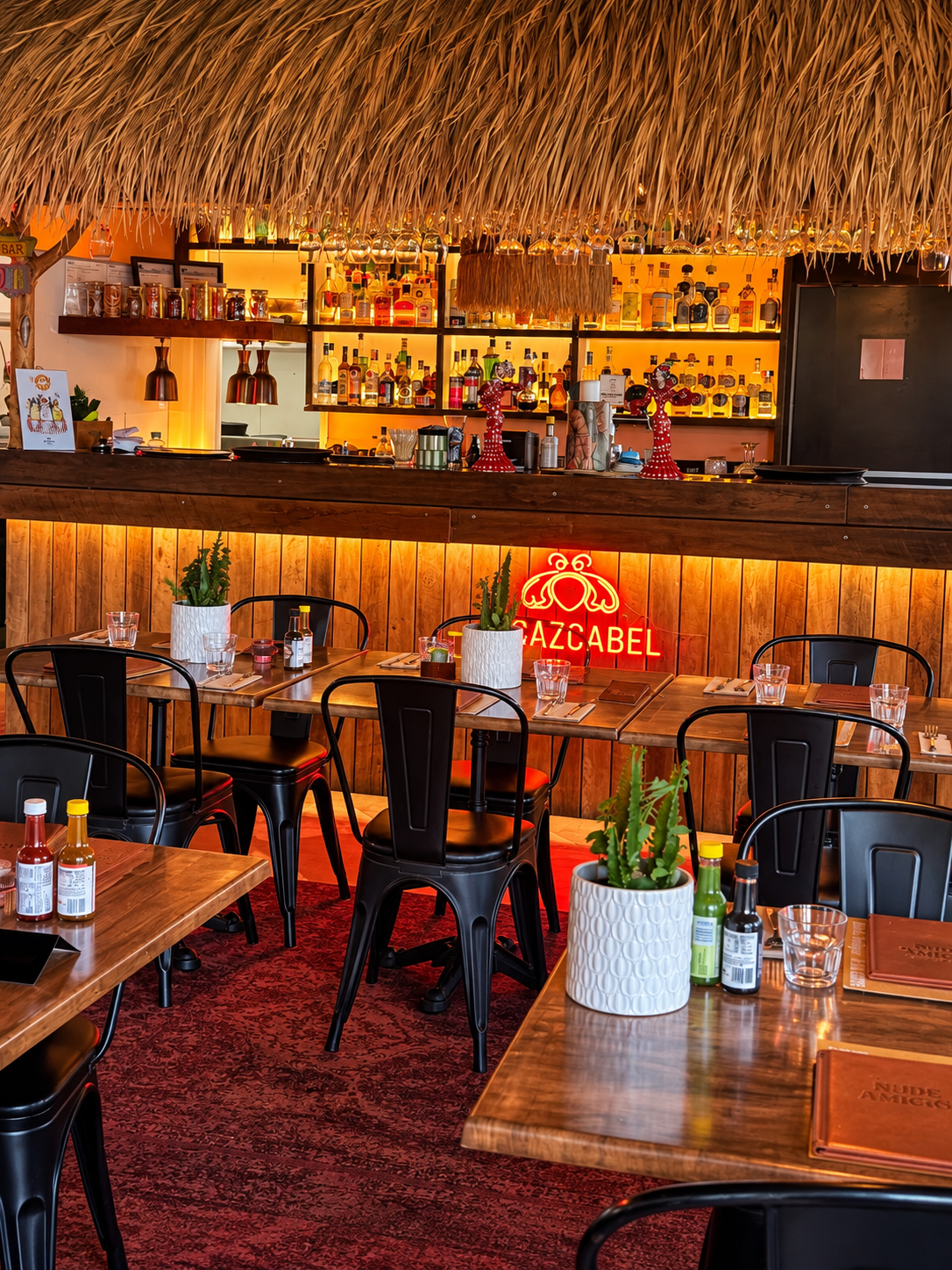 Interior of a restaurant with wooden tables, black chairs, potted plants, and a bar area with bottles of alcohol and festive decorations.